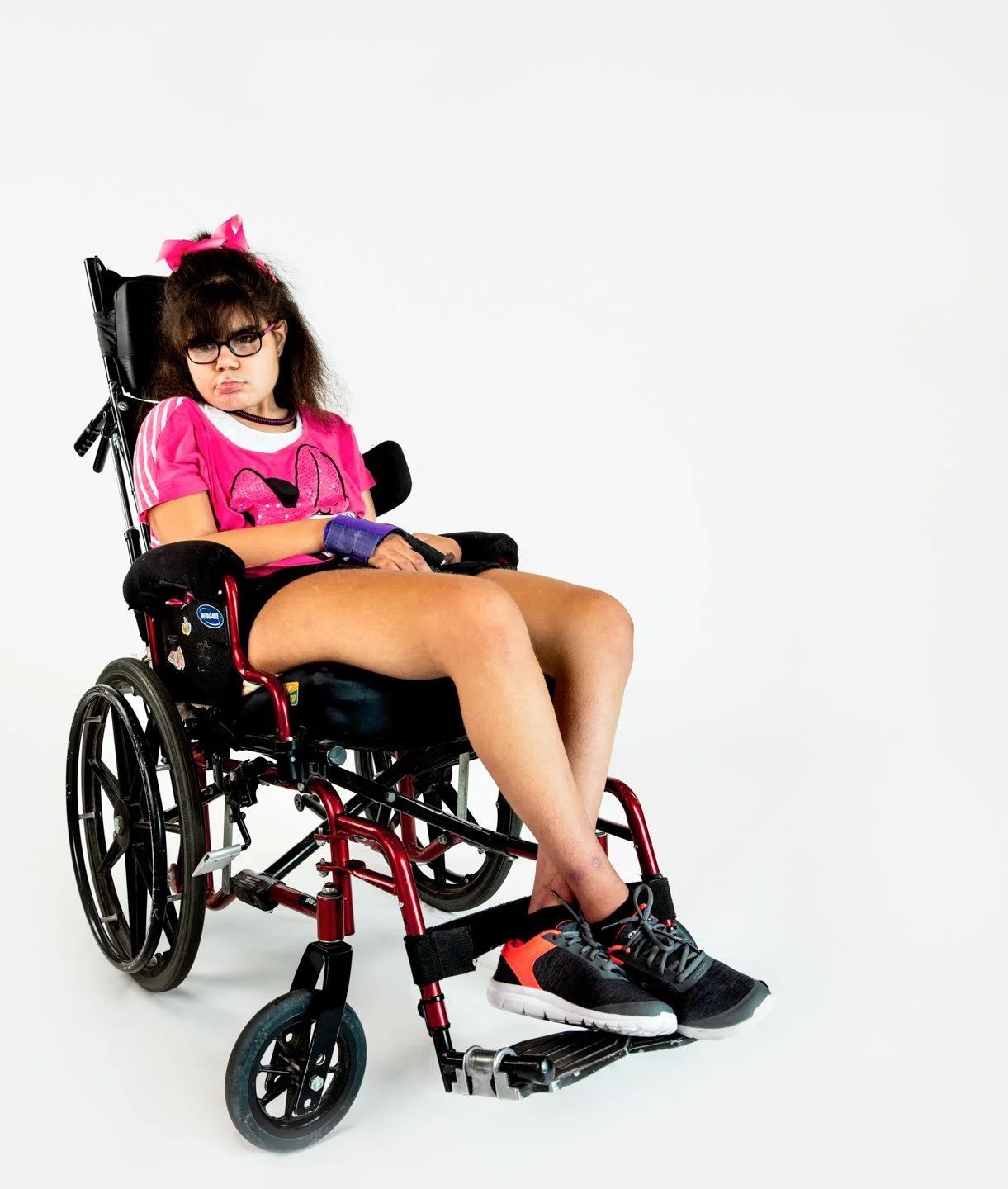 Young woman with dark curly hair in a wheelchair, wearing a pink t-shirt, black shorts, black and red sneakers, and glasses, sitting against a plain white background.