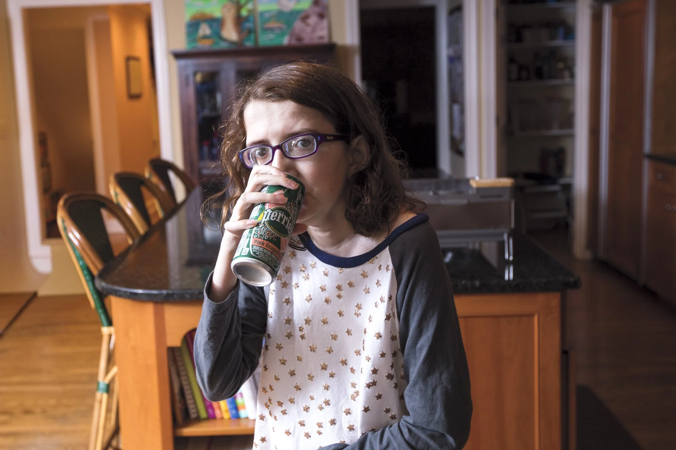 A young girl with curly brown hair and glasses drinks from a can of Pibb Xtra cola in a kitchen. She wears a white shirt with gold stars and gray sleeves. The background shows wooden cabinets, a black countertop, and kitchen chairs.