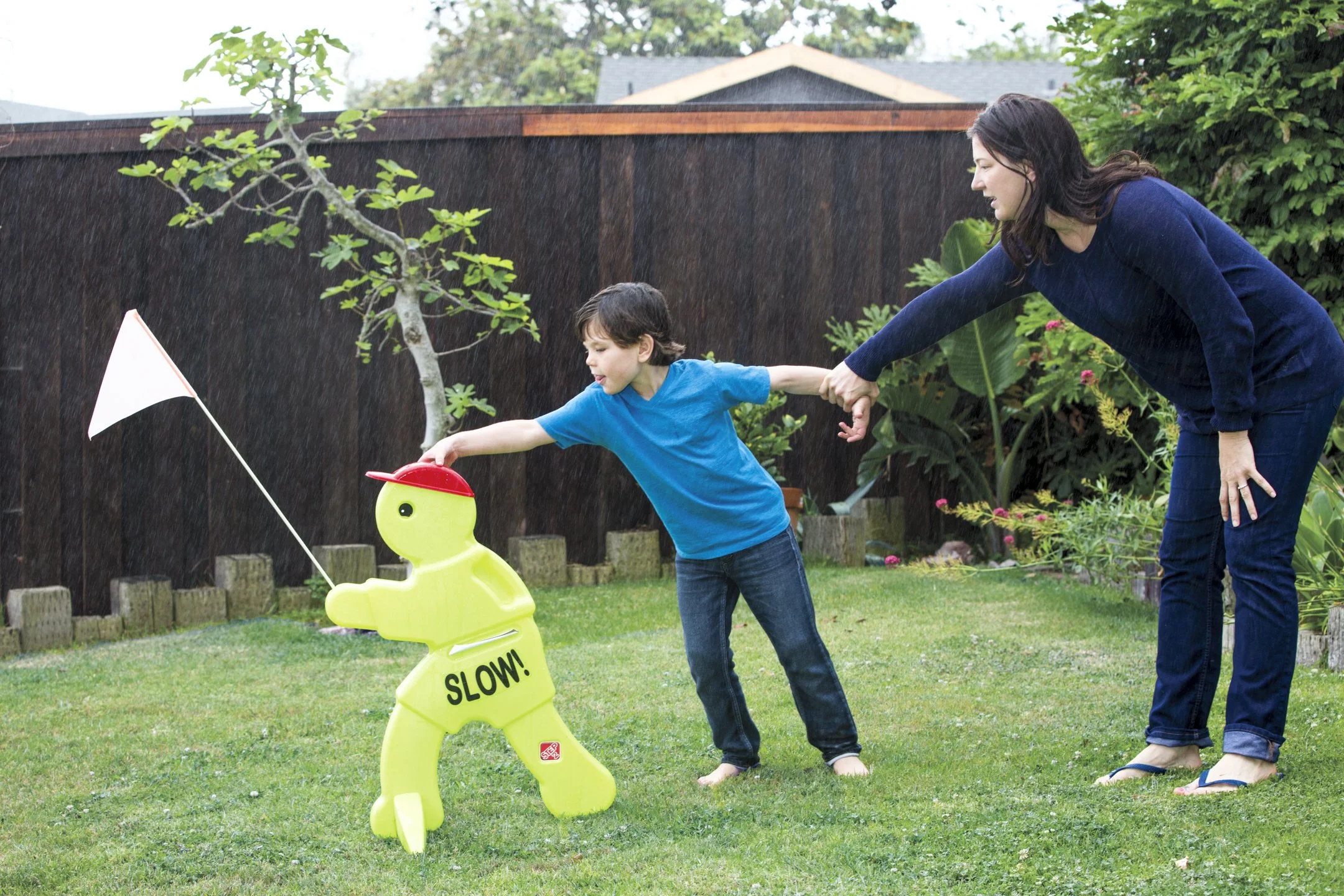 A woman and a boy playing tug-of-war with a lawn sprinkler toy labeled 'SLOW!' in a backyard during rain.