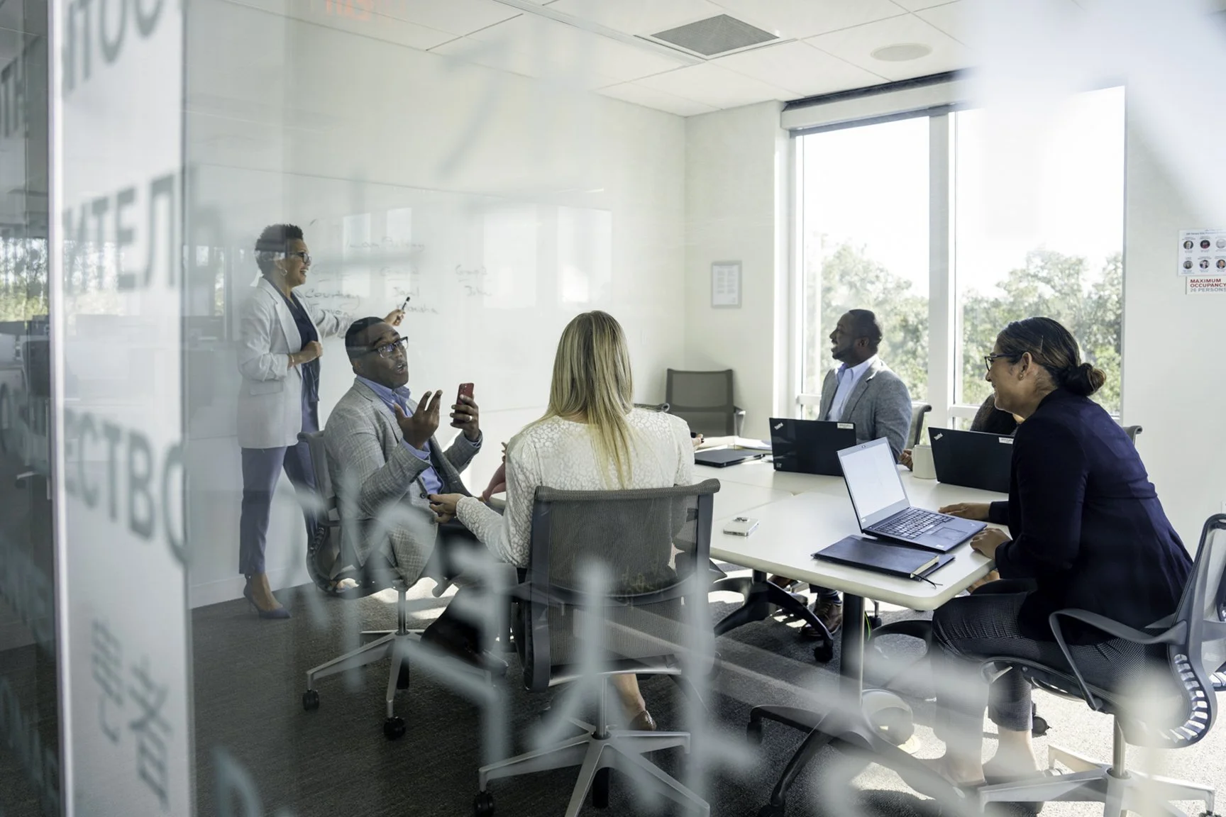 Business meeting in a modern conference room with five people, four seated at a table with laptops, one standing and presenting at a whiteboard, seen through a glass wall.