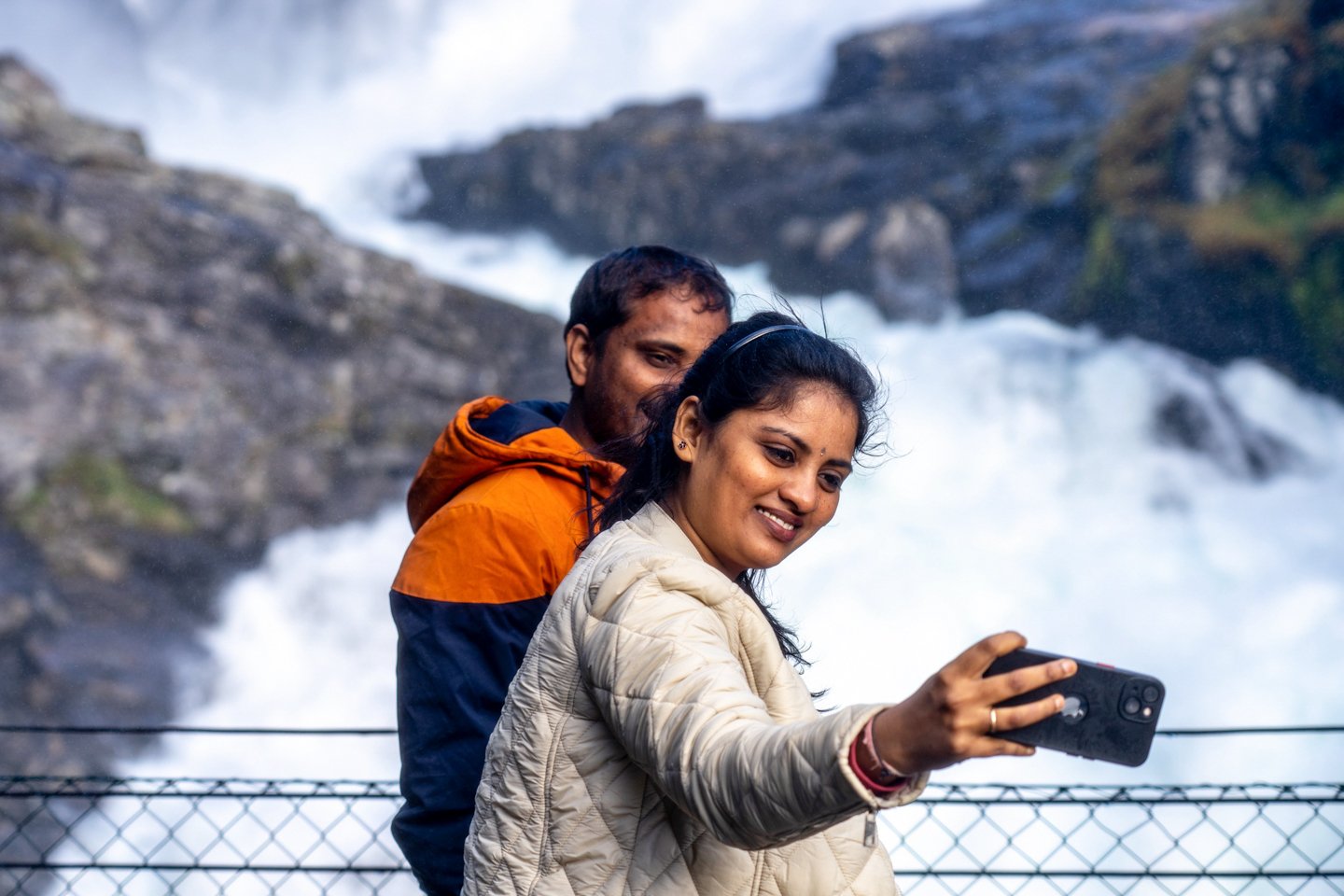 A woman and a man stand together outdoors near a waterfall, with the woman taking a photo with her phone.
