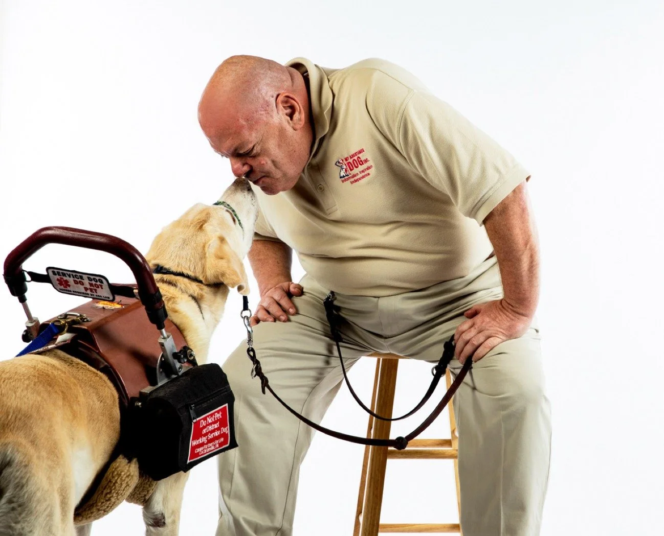 A man in a beige uniform sitting on a stool and touching noses with a service dog that is wearing a vest labeled 'Do Not Pet' and 'Service Dog'. The man appears to be a trainer or handler, inside a studio with a plain white background.