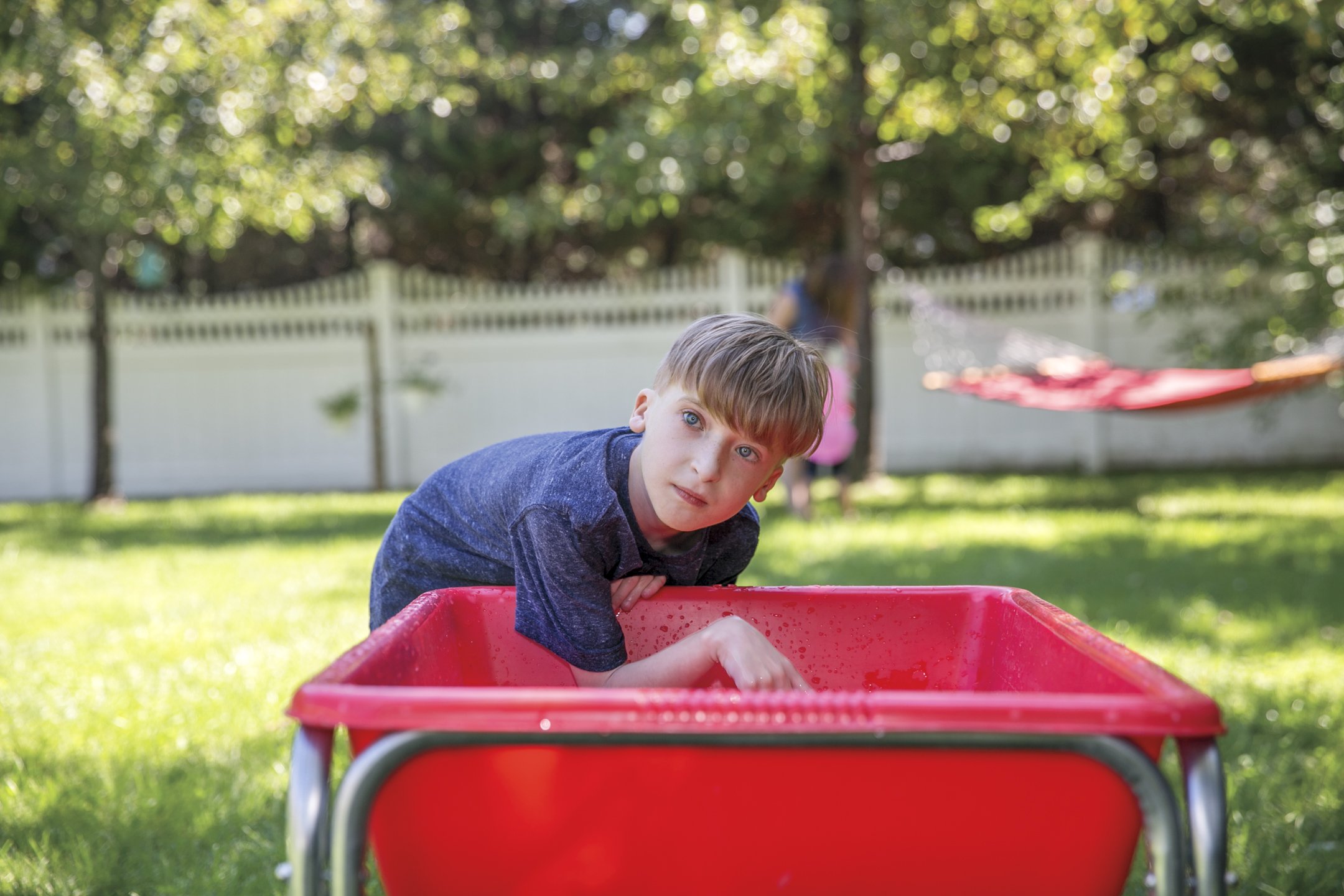 A young boy with blond hair and blue eyes leaning over a red wheelbarrow in a backyard with green grass, trees, and a white fence. A girl in pink is blurred in the background near a hammock.
