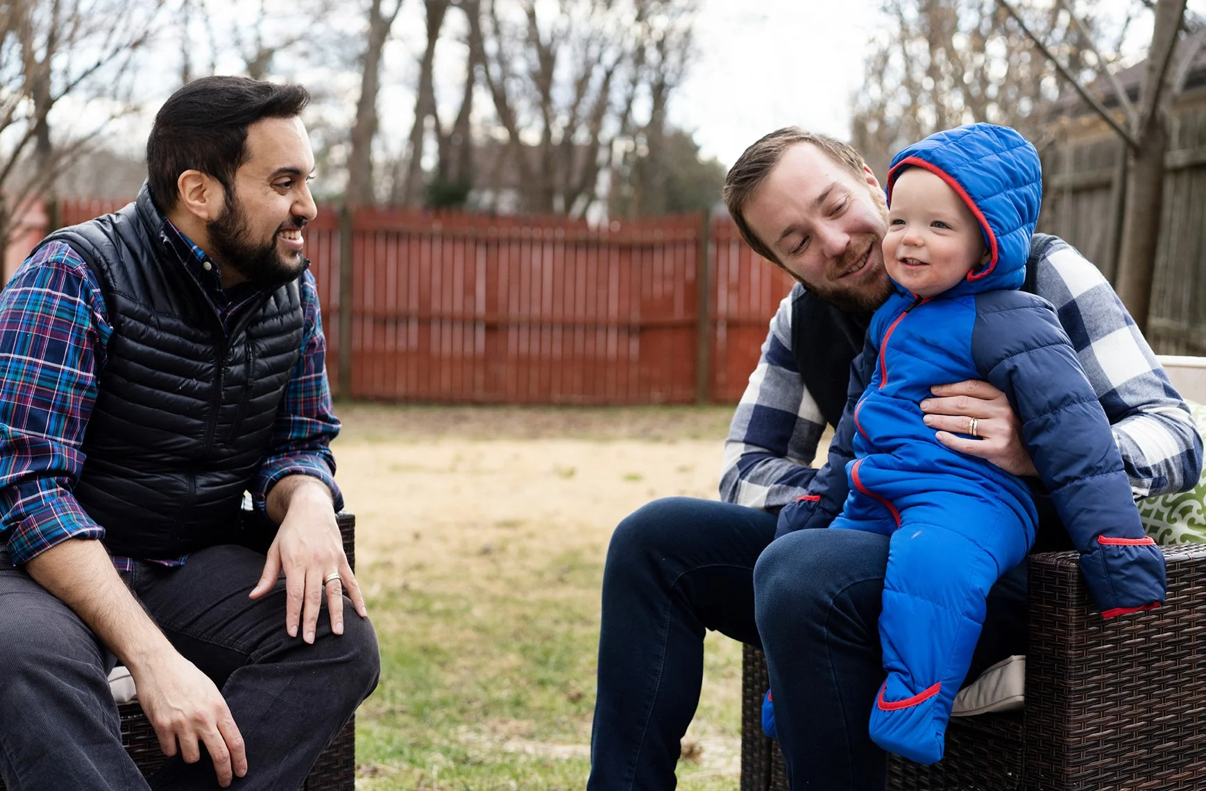 Two men and a toddler sitting outside in a yard with a brown fence in the background. One man is sitting on a chair, smiling, while the other man is holding and playing with the toddler on his lap. The toddler is dressed in a blue winter jacket with 