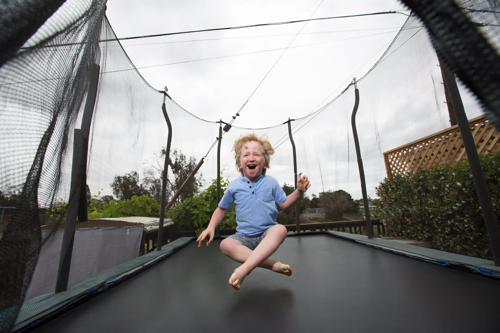 Child jumping on a trampoline with a safety net on a cloudy day in a backyard.