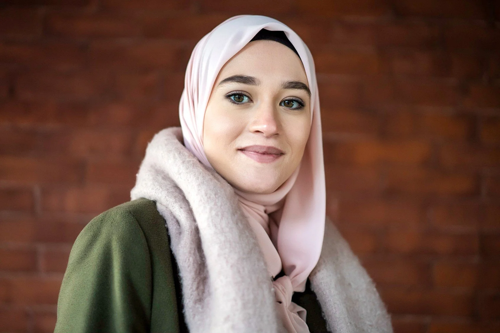 A woman wearing a pink hijab and a green coat standing in front of a brick wall.