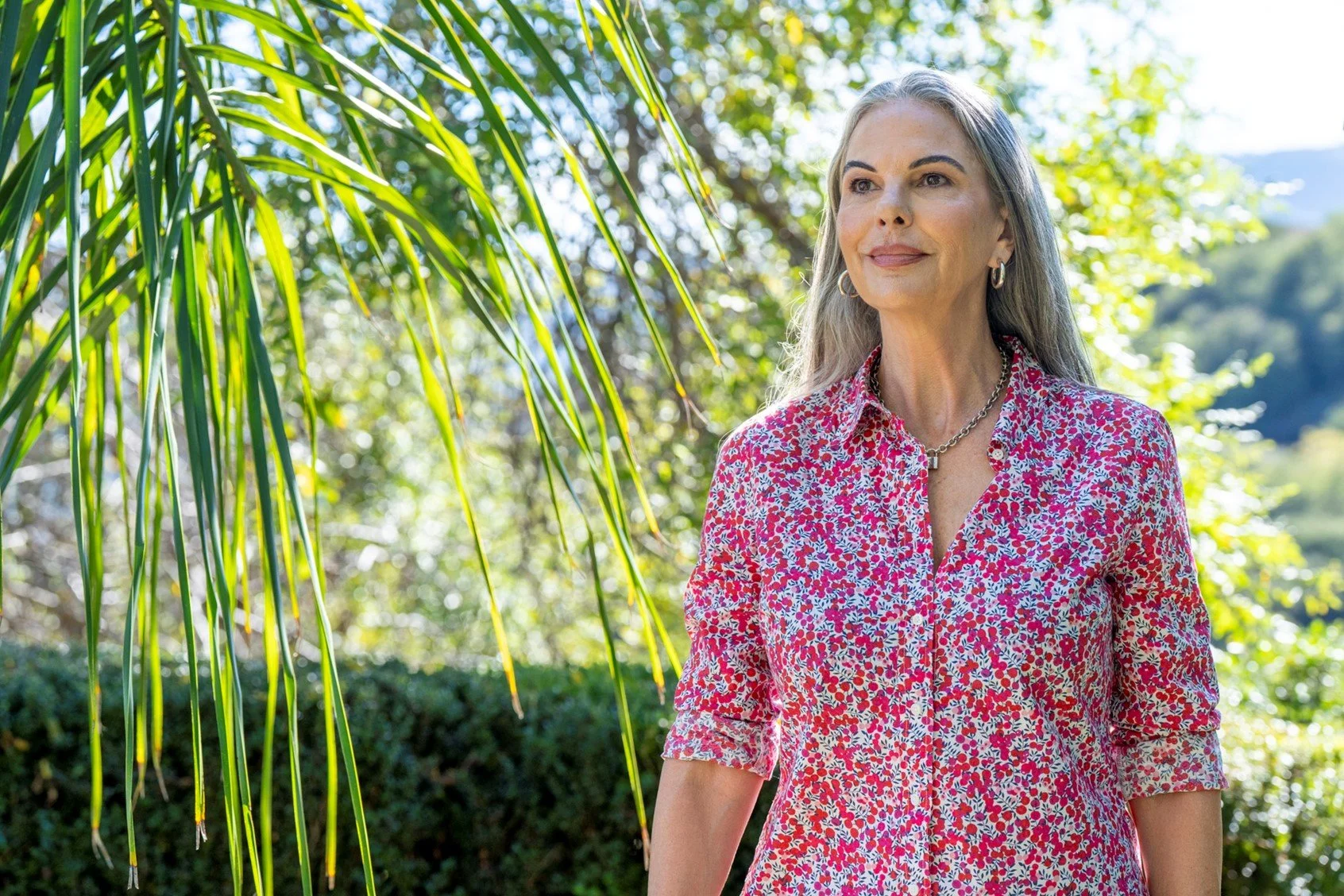 A woman with gray hair and hoop earrings standing outdoors among green trees and foliage, wearing a red and pink floral blouse. Sunlight filters through the leaves.