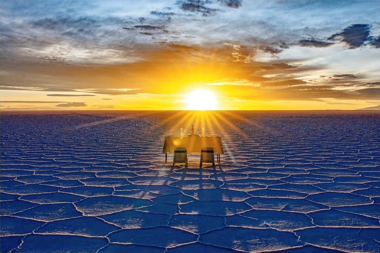 A desert scene with a table and two chairs set up on salt flats during sunset, with a vibrant sky and clouds in the background.