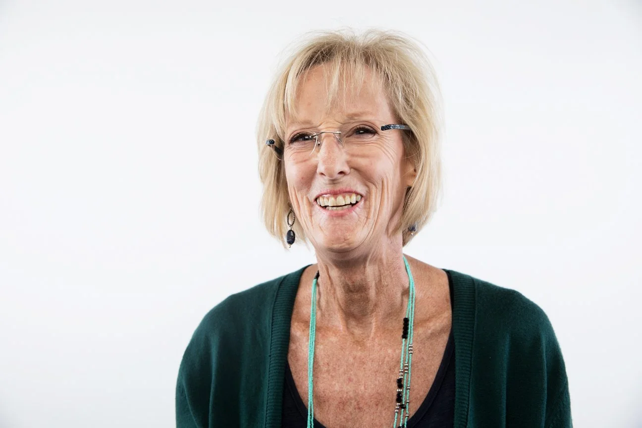 A smiling elderly woman with short blonde hair, wearing glasses, a dark green top, and a beaded necklace, standing against a plain white background.