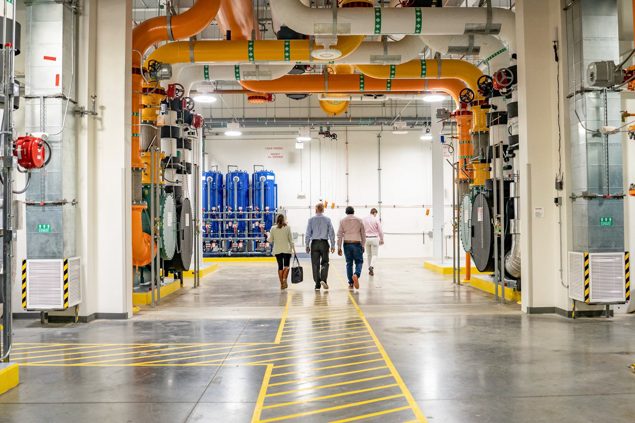 Four people walking through an industrial facility with large orange and white pipes overhead and blue equipment in the background.