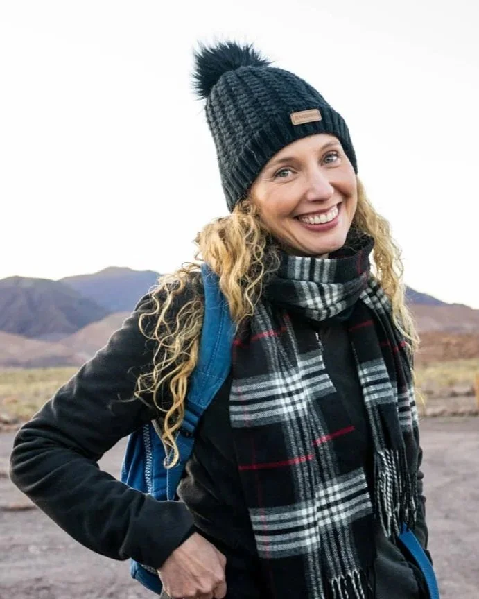 Smiling woman in outdoor winter gear, including a black and gray beanie with a pom-pom, a plaid scarf, and a blue backpack, standing in a mountainous desert landscape.