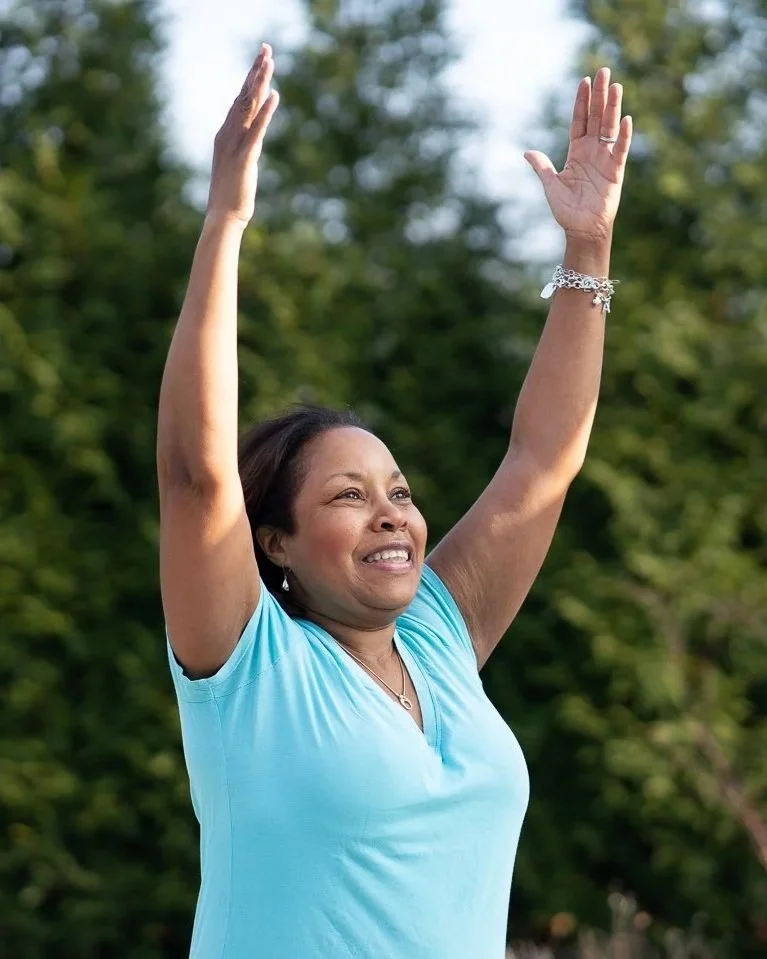 A woman in a blue shirt raising her arms with a smile outdoors, with green trees in the background.