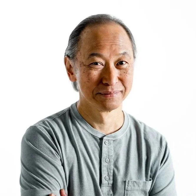 A middle-aged Asian man with gray hair, wearing a gray short-sleeve shirt, standing with arms crossed against a plain white background.
