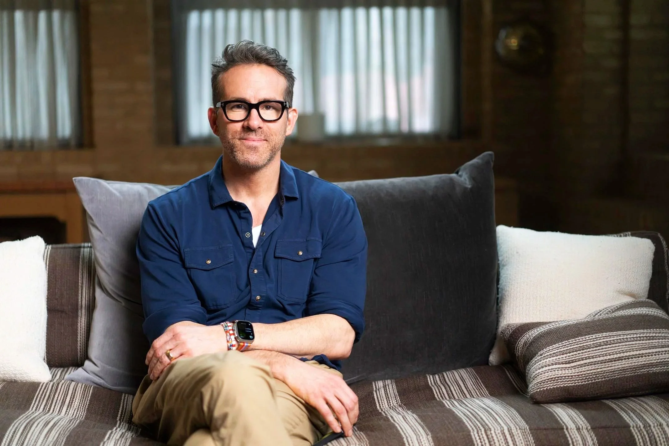 A man with glasses, gray hair, and a beard sitting on a sofa in a cozy living room, wearing a blue button-up shirt, beige pants, and a smartwatch.