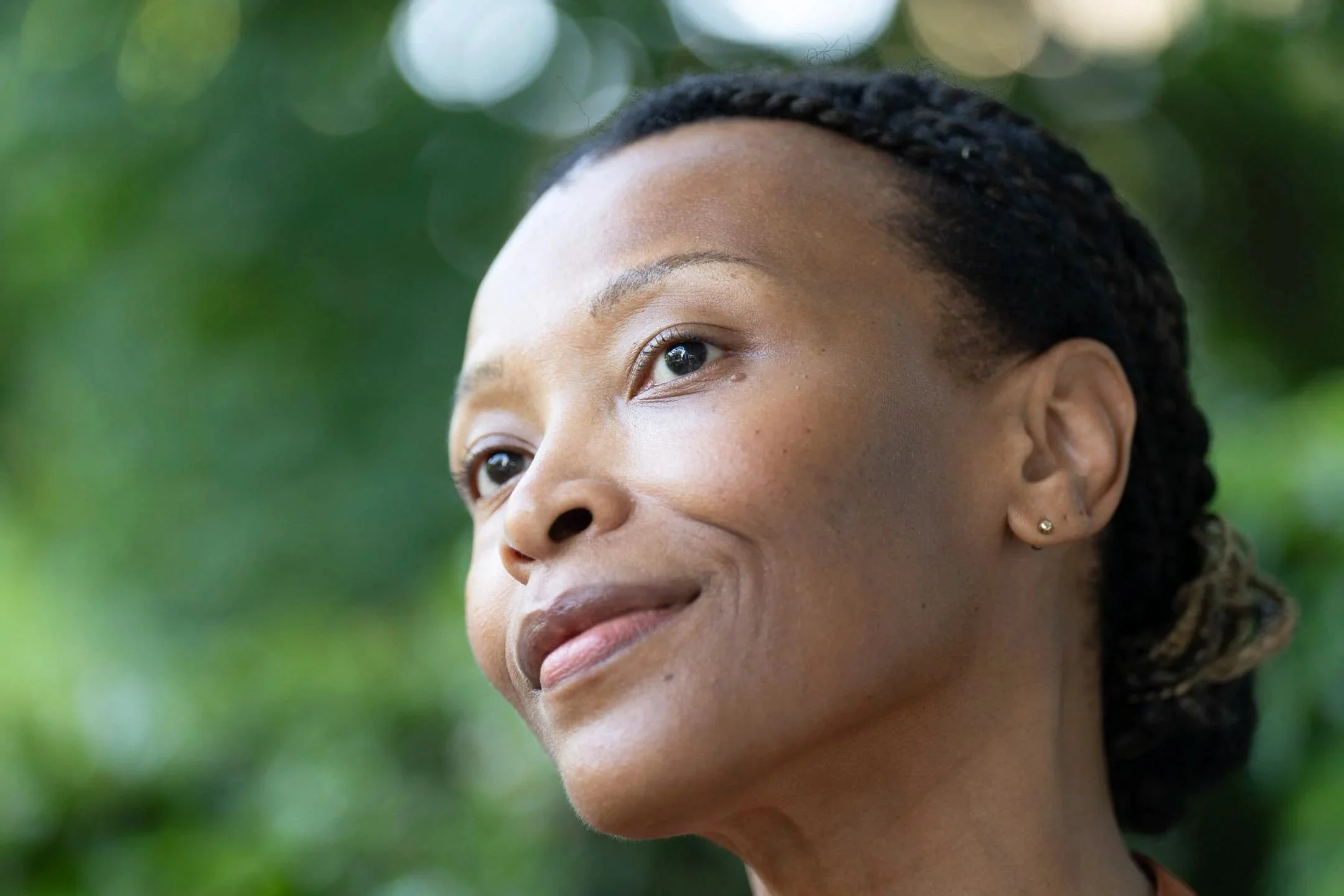 Close-up of a woman with a calm expression, looking to the side outdoors with a blurred green background. Beauty campaign, Strivectin