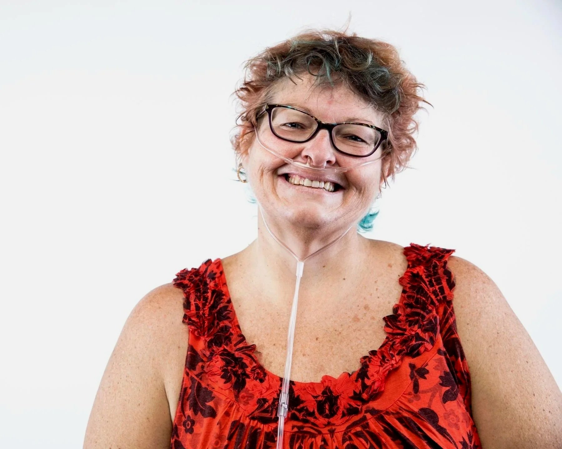 Smiling woman with medical tube in nose, wearing glasses and a red floral sleeveless top, against a plain white background.