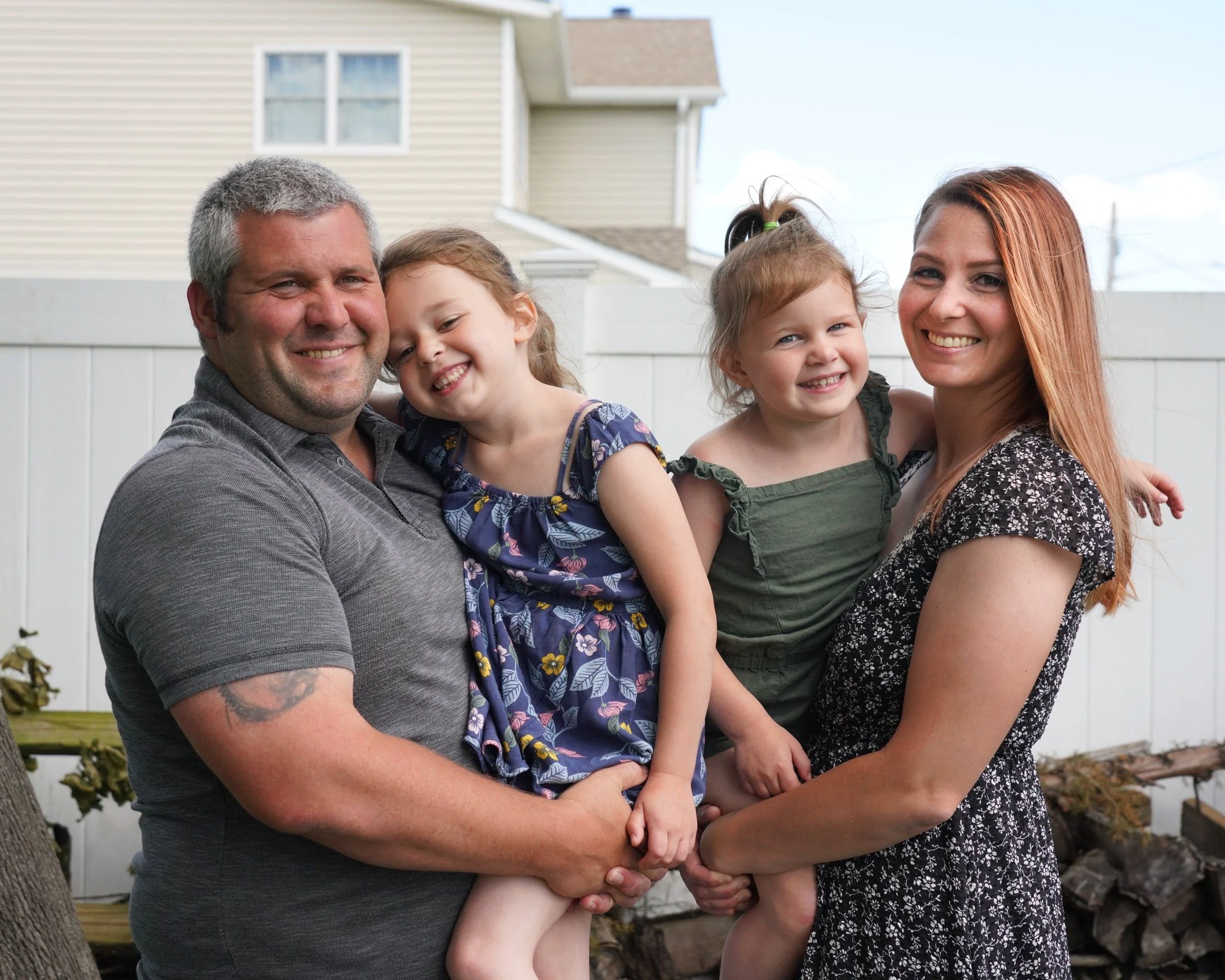 A smiling family of four outdoors in a backyard, with a man and woman holding two young girls, all smiling and looking at the camera.