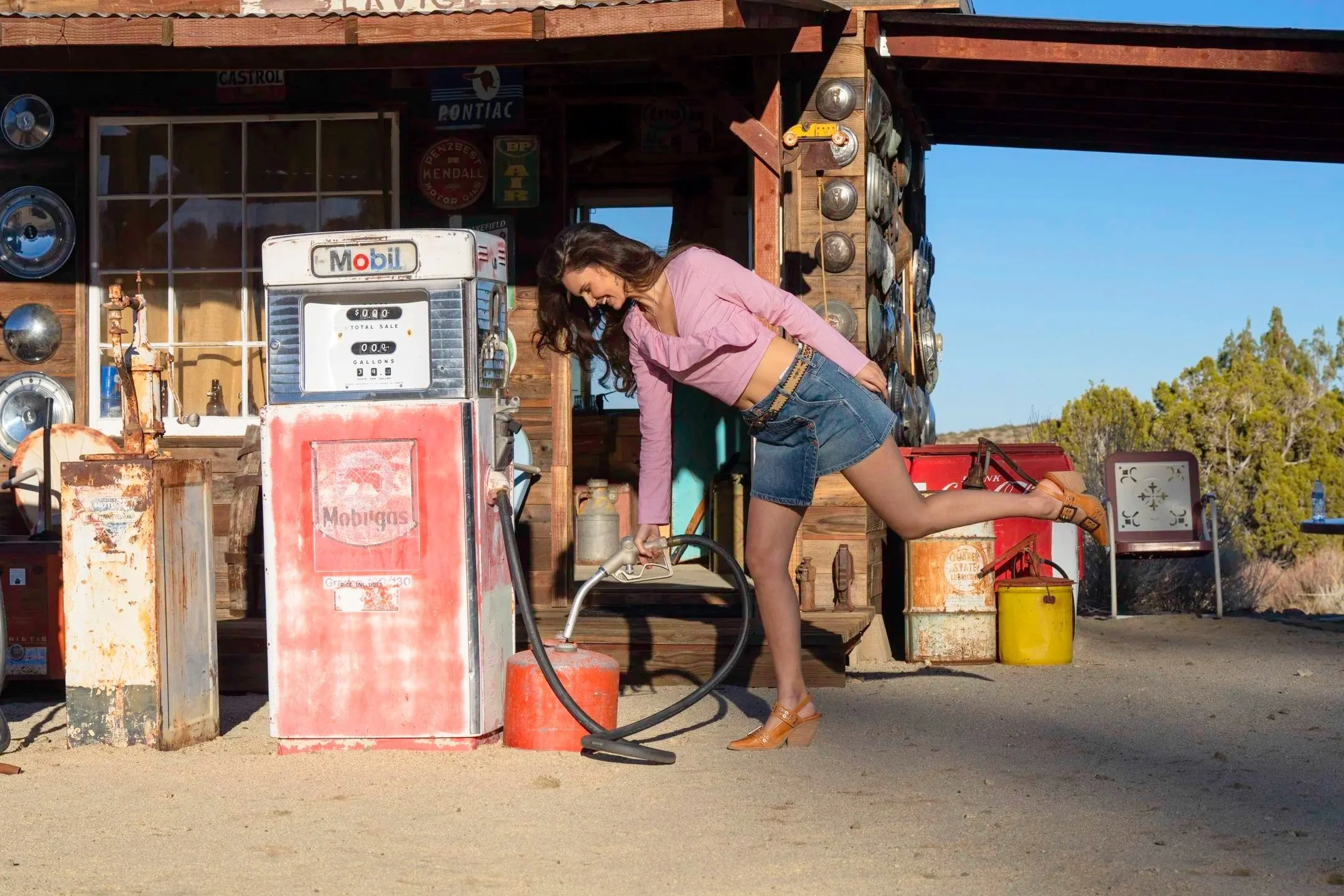 A woman in a pink shirt and denim skirt leaning over an old gas pump at a rustic outdoor gas station, holding the pump hose with one hand and lifting her foot with the other, surrounded by vintage signs and equipment.