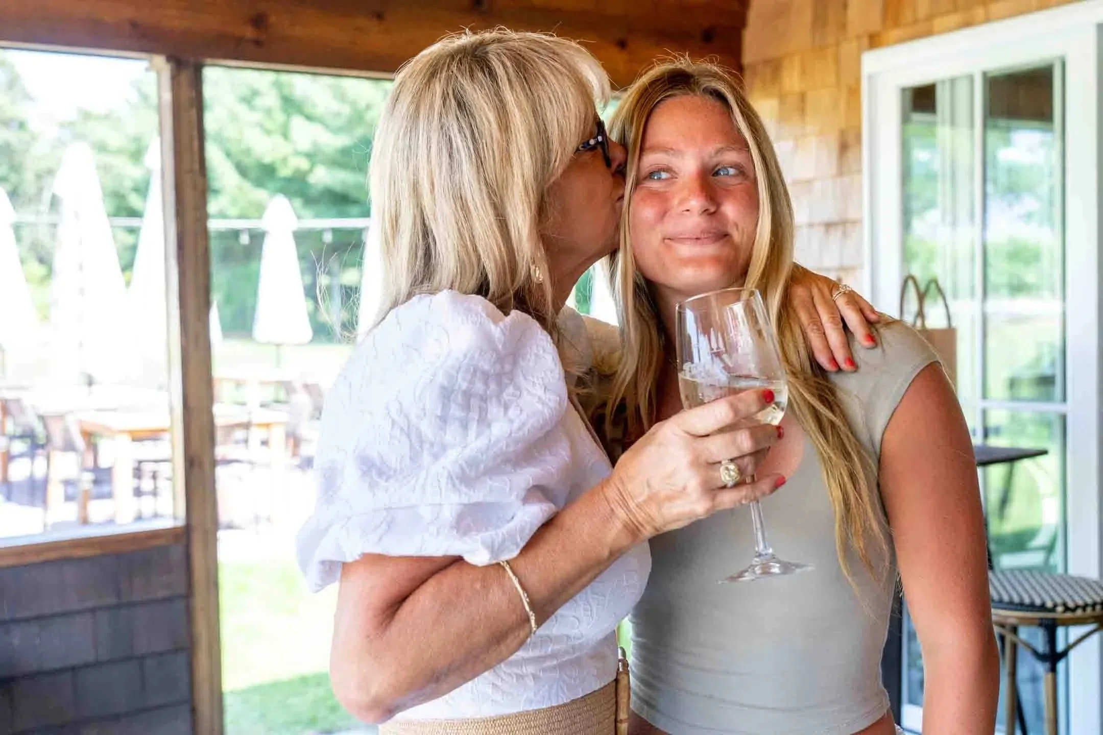 An older woman is kissing a young woman on the cheek while holding a glass of white wine. The young woman is smiling as she looks slightly to the side. They are inside a bright room with large windows and outdoor greenery visible in the background.