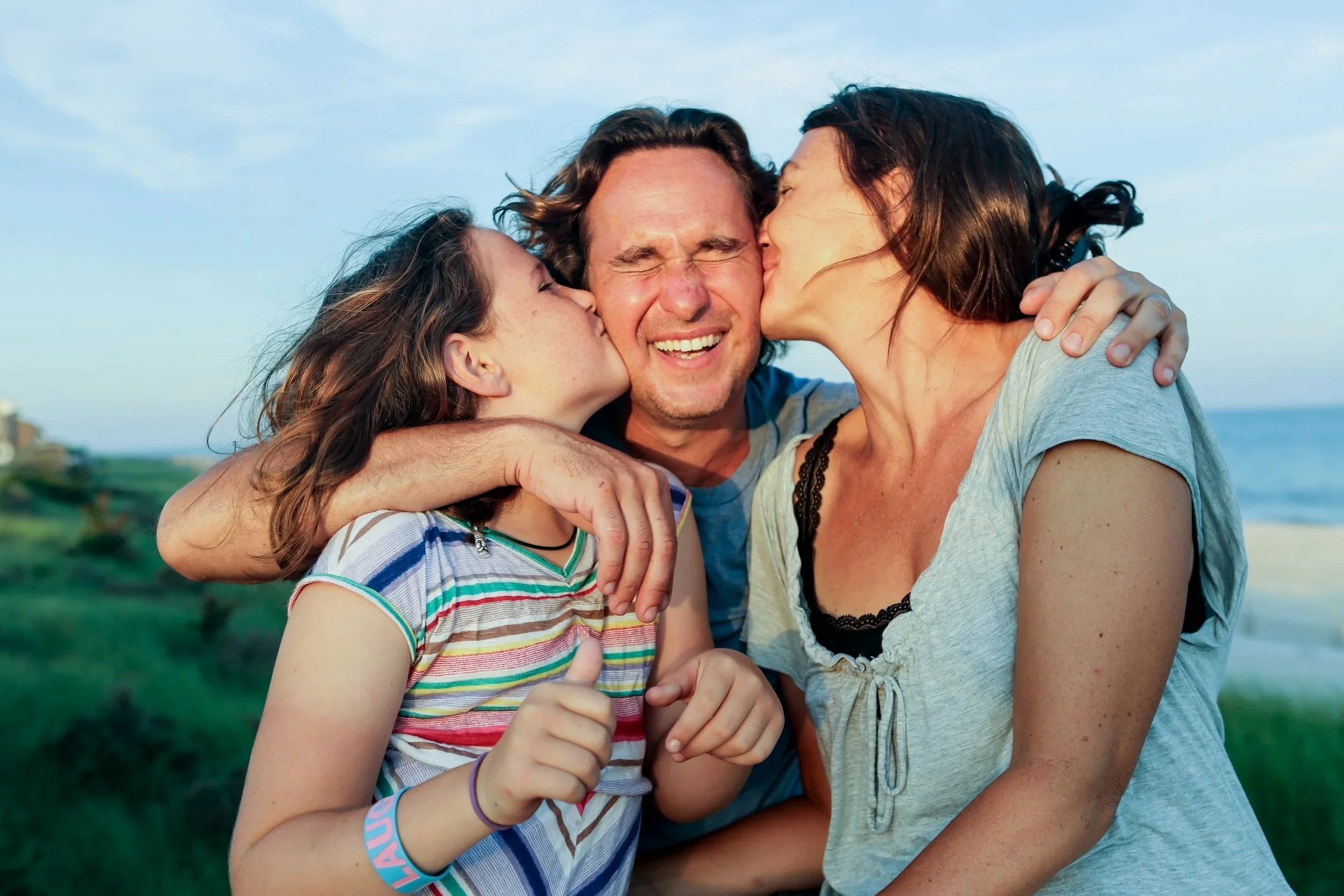 A man being kissed on both cheeks by two women outdoors near the coast.