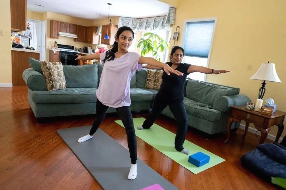 Two women practicing yoga indoors, each on a yoga mat with their arms extended forward, in a living room area with a couch, side table, and kitchen in the background.