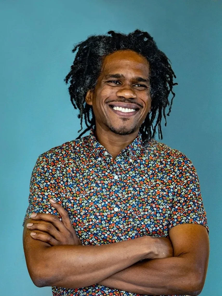 A man with dreadlocks smiling with his arms crossed, wearing a colorful floral shirt, standing against a blue background.