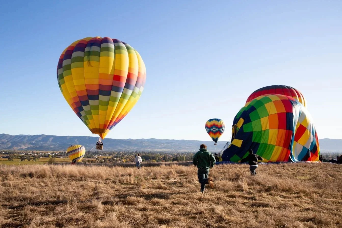 Several colorful hot air balloons are being prepared for takeoff in an open field with dry grass. Some balloons are partially inflated on the ground, while others are floating in the air, with mountains and a clear blue sky in the background.