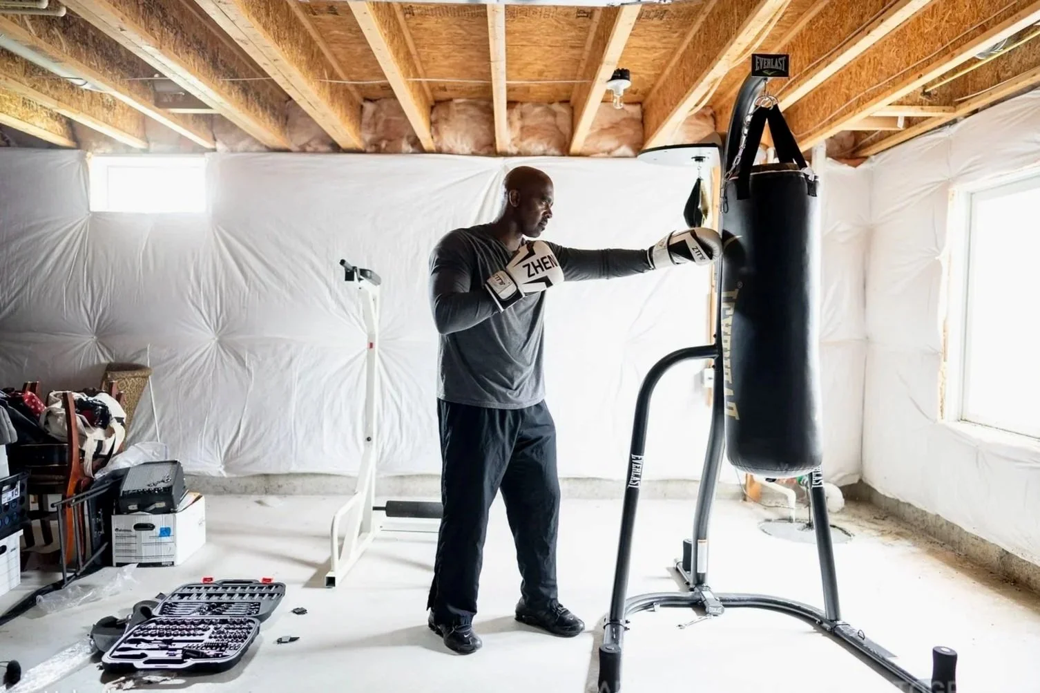 A man wearing boxing gloves practicing punches on a heavy punching bag in a basement gym. The room is unfinished, with exposed wooden beams and white insulation on the walls. There are boxes and a toolbox on the floor, and a treadmill behind him.