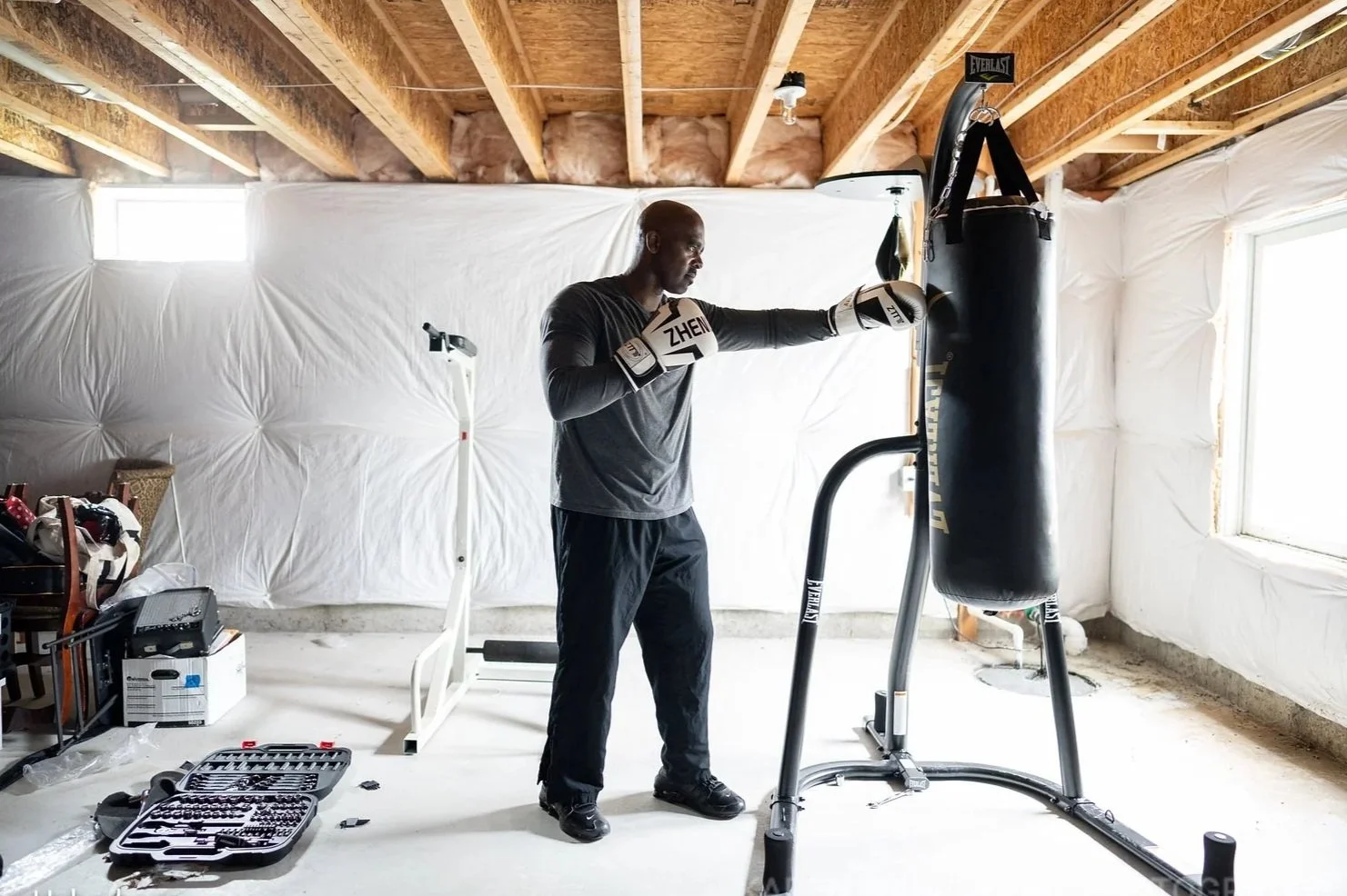 A man wearing boxing gloves practicing punches on a heavy punching bag in a basement gym. The room is unfinished, with exposed wooden beams and white insulation on the walls. There are boxes and a toolbox on the floor, and a treadmill behind him.