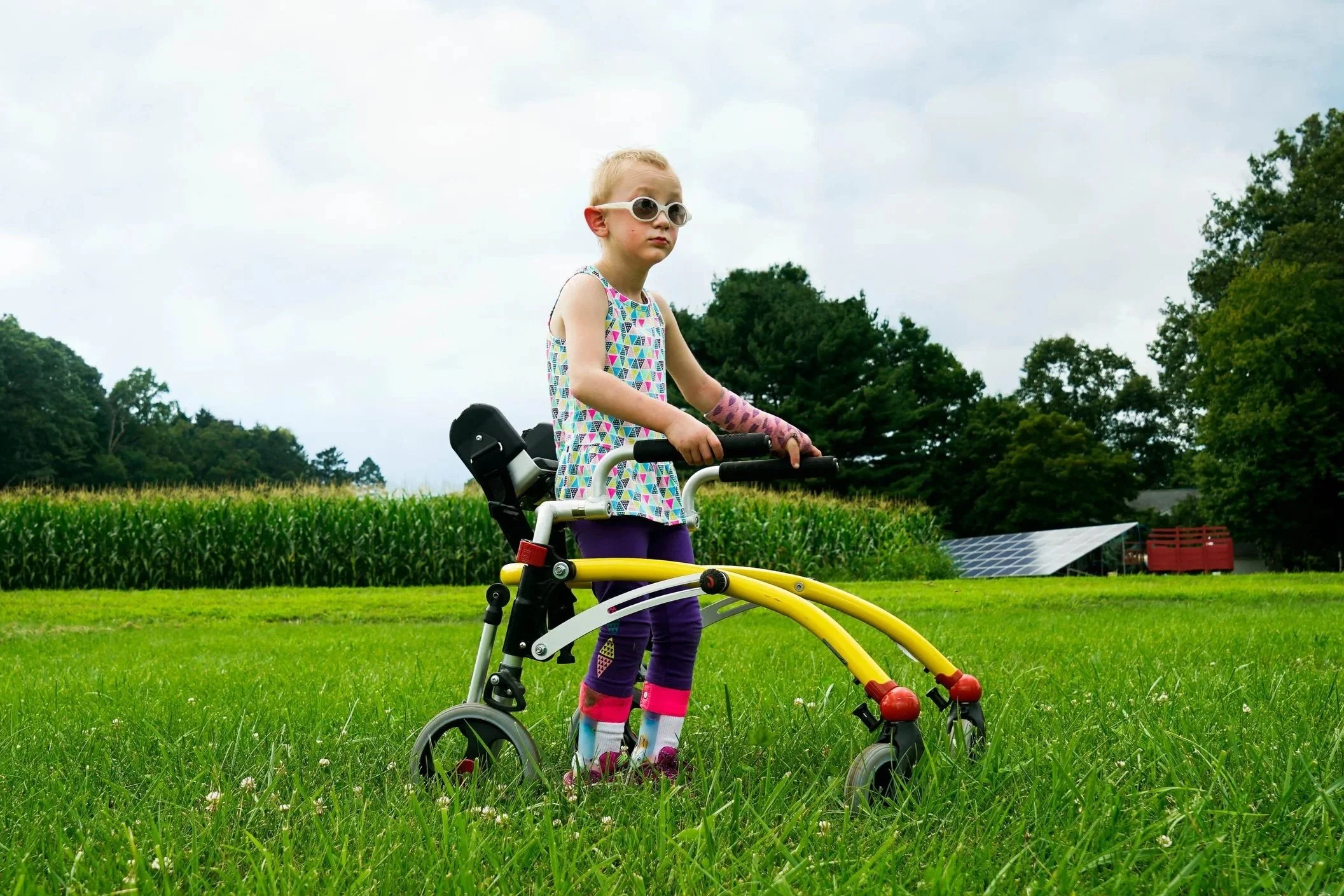 A young girl with sunglasses and colorful clothing standing on a walker outdoor on a grassy field with trees and a farm in the background.