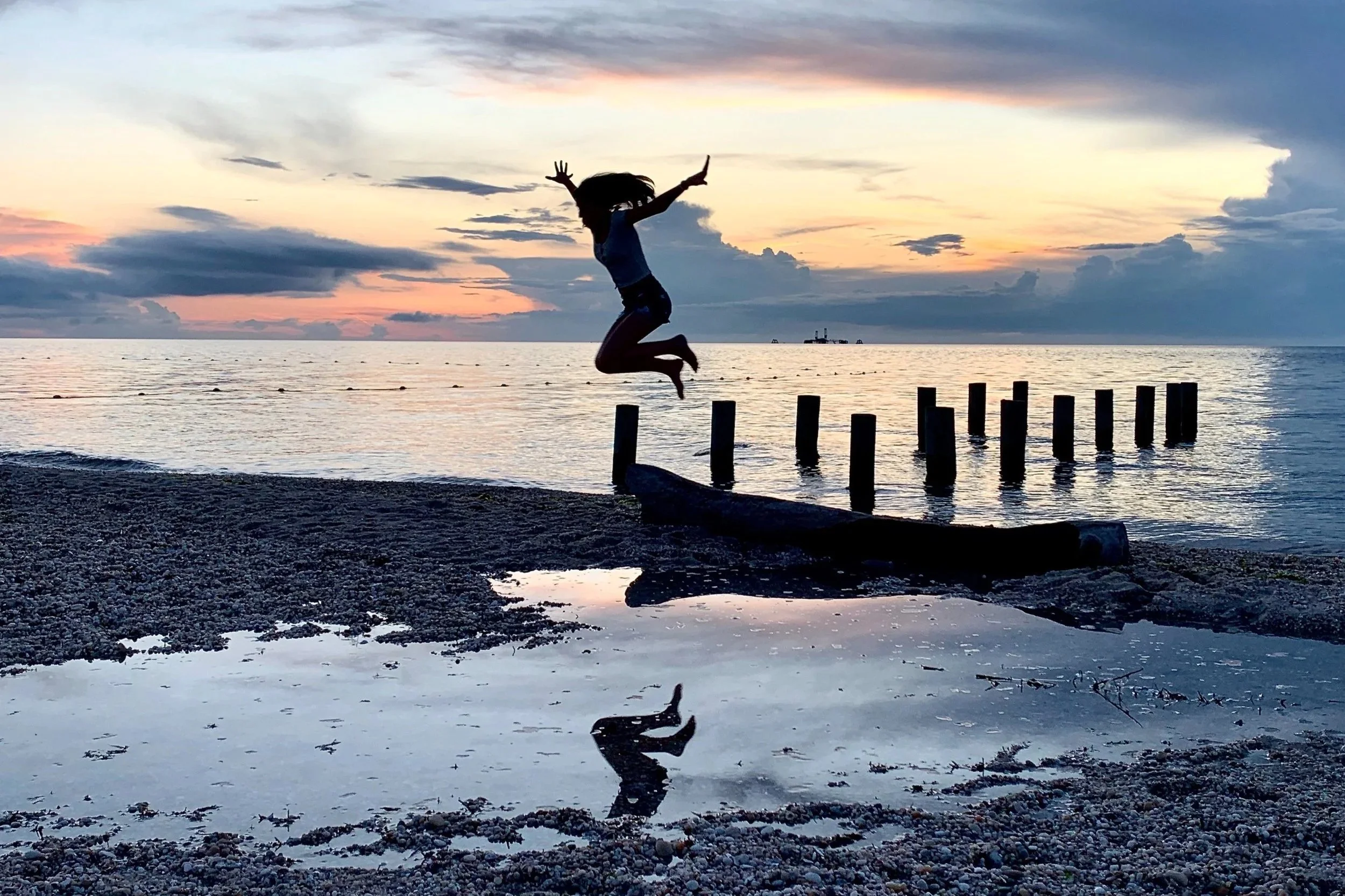 Person jumping on a pebble beach at sunset, with their reflection in a small pool of water, calm ocean, cloudy sky, and wooden posts in the background.