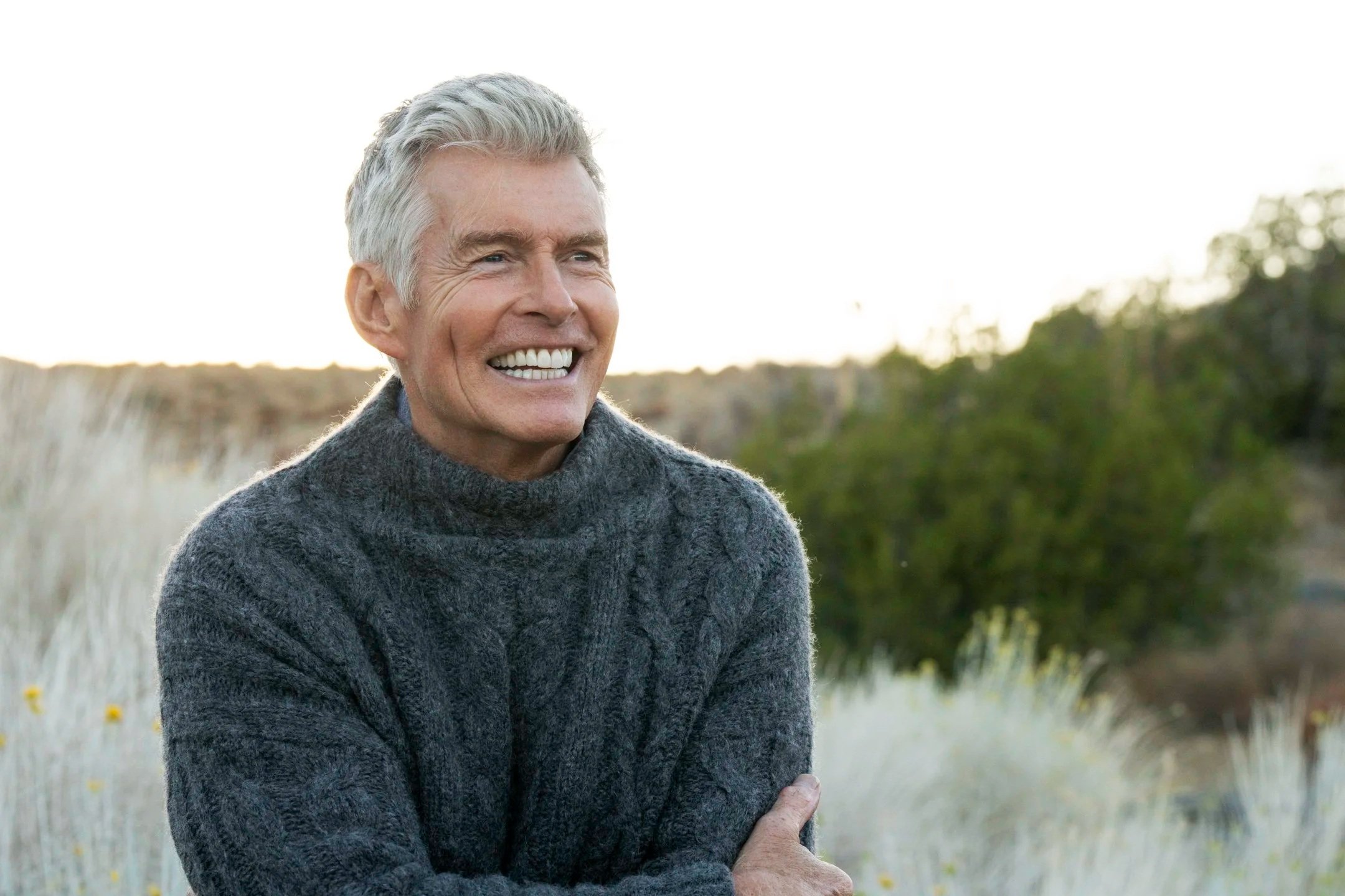 An elderly man with gray hair and a wide smile wearing a dark gray knitted sweater outdoors in a natural landscape.
