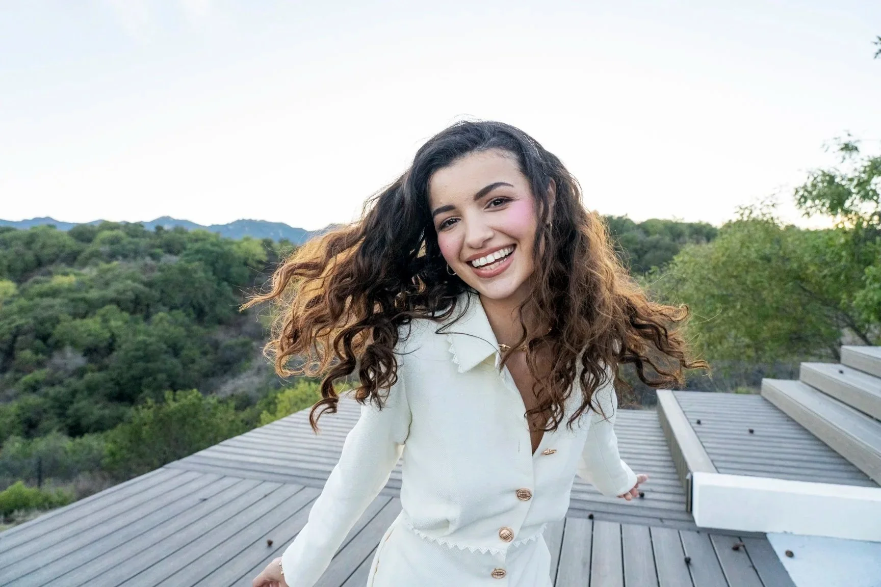 A young woman with long, curly brown hair smiling and leaning on a wooden deck railing outdoors with green trees and distant mountains in the background.