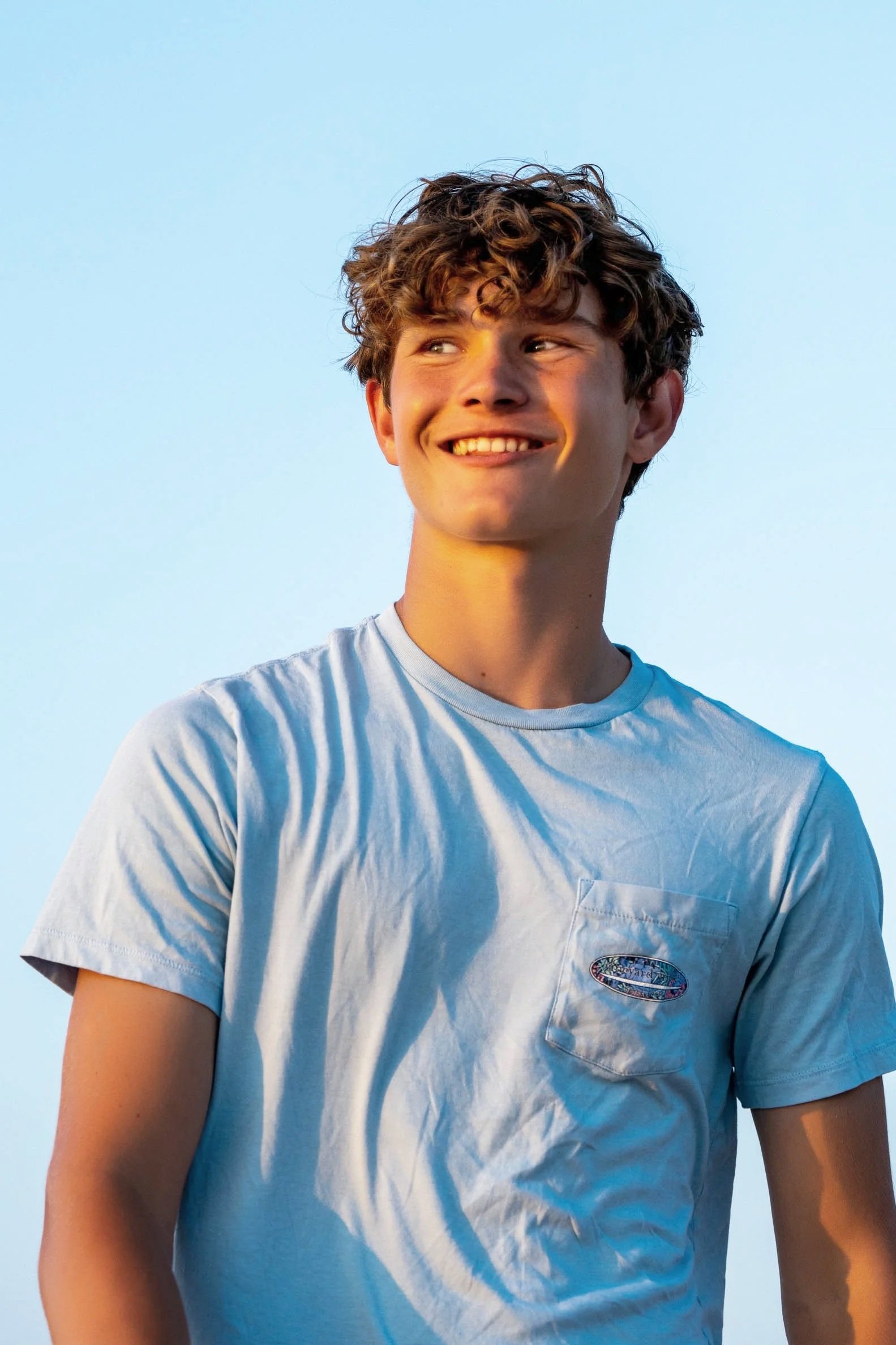 A young man with curly brown hair smiling and looking to the side, wearing a light blue t-shirt with a small pocket and a colorful patch, standing outdoors against a clear blue sky.