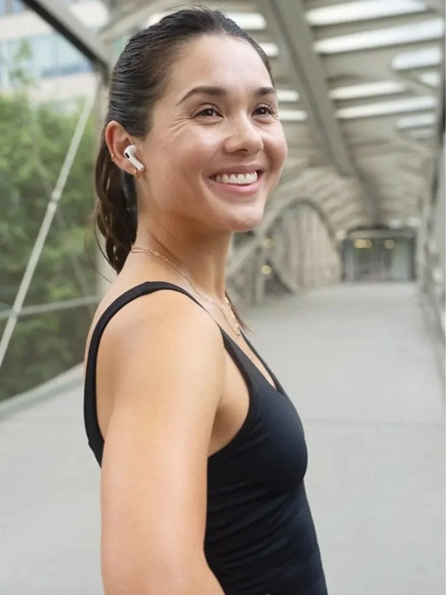 Smiling woman wearing a black athletic tank top, wireless earbuds, and a gold necklace in a modern covered outdoor walkway.