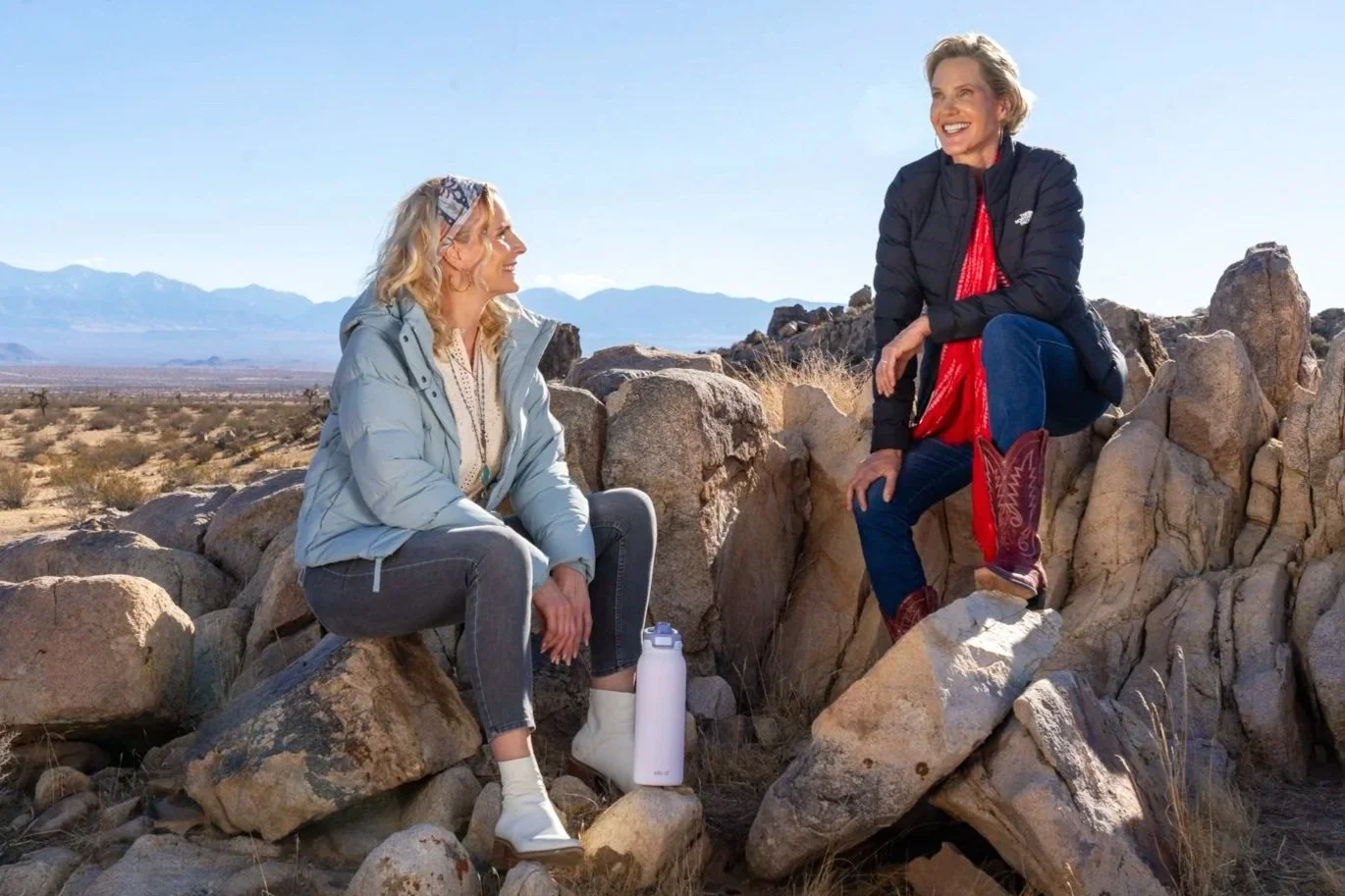 Two women sitting and talking on rocks in a desert landscape with mountains in the background, sunny weather, one wearing a light jacket and the other a black jacket with cowboy boots.
