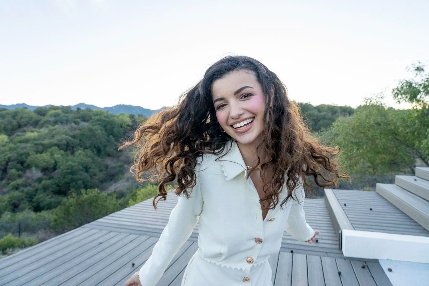A young woman with long, curly hair smiling on a rooftop with mountains and trees in the background.