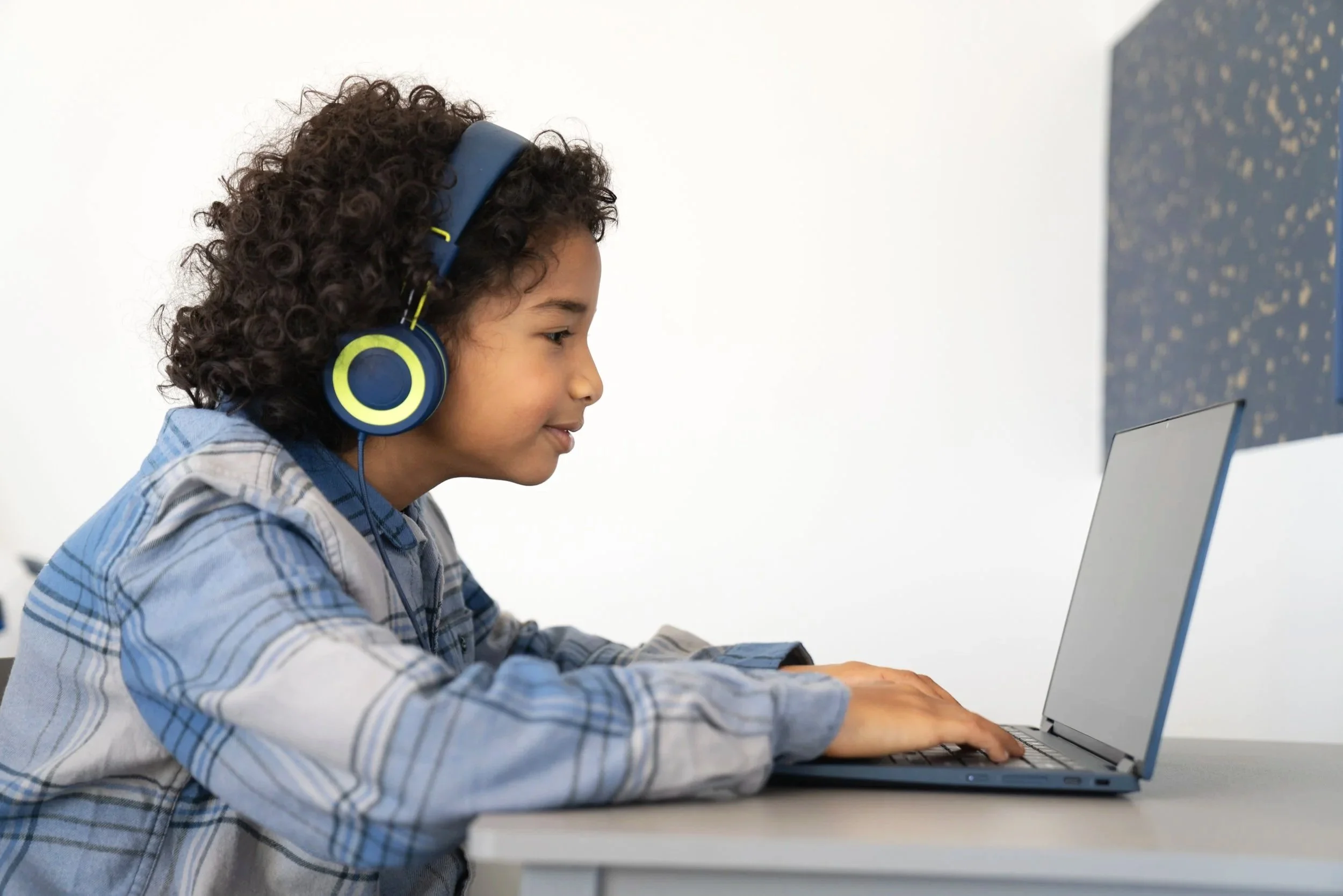 A young boy with curly hair wearing headphones and a blue plaid shirt, sitting at a desk typing on a laptop. The background is plain white with a dark patterned wall to the right.