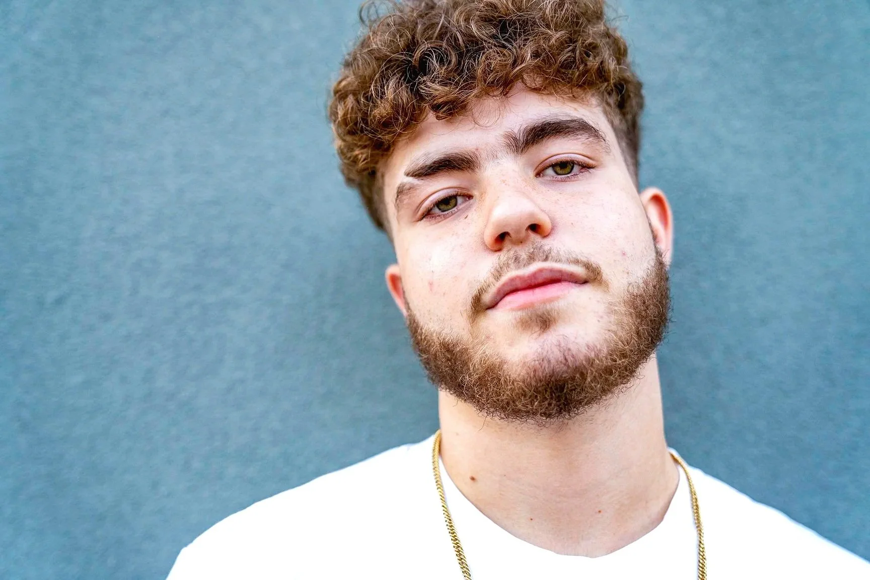 Close-up of a young man with curly hair, beard, wearing a white shirt and gold chain, standing against a blue background.