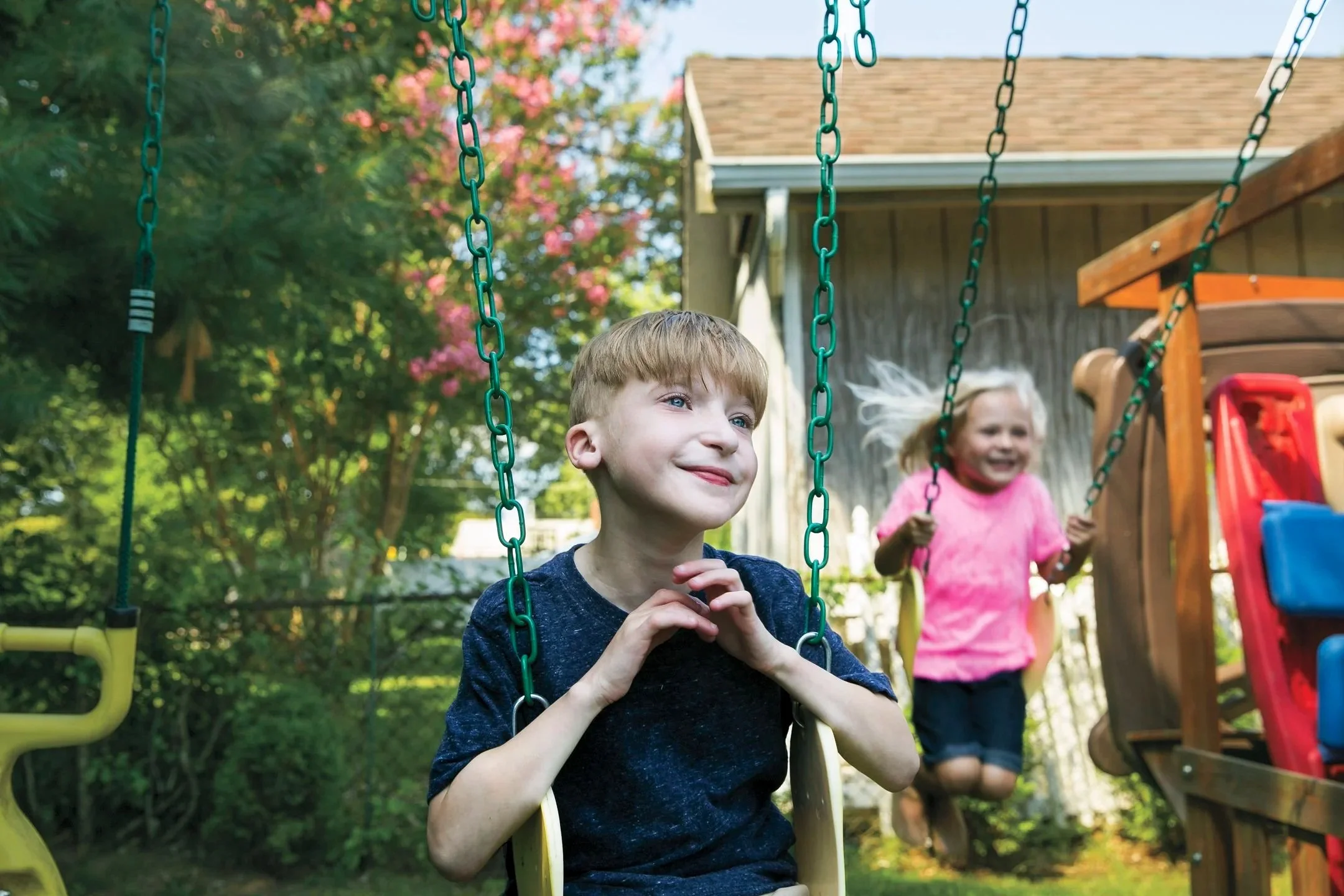 Two children, a boy and a girl, playing on swings in a backyard with trees and a shed.