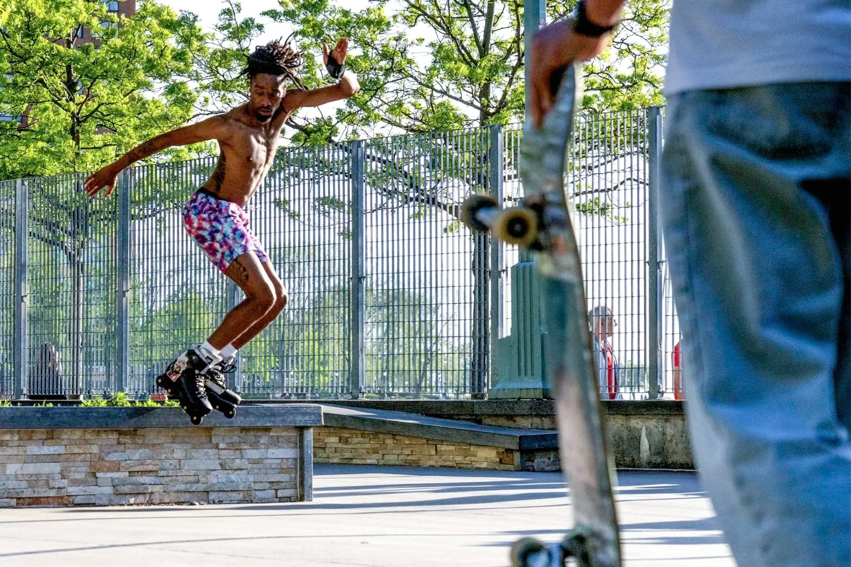 A young man is rollerblading on a low ledge at a skatepark while another person holds a skateboard nearby. There is green foliage and a metal fence in the background.