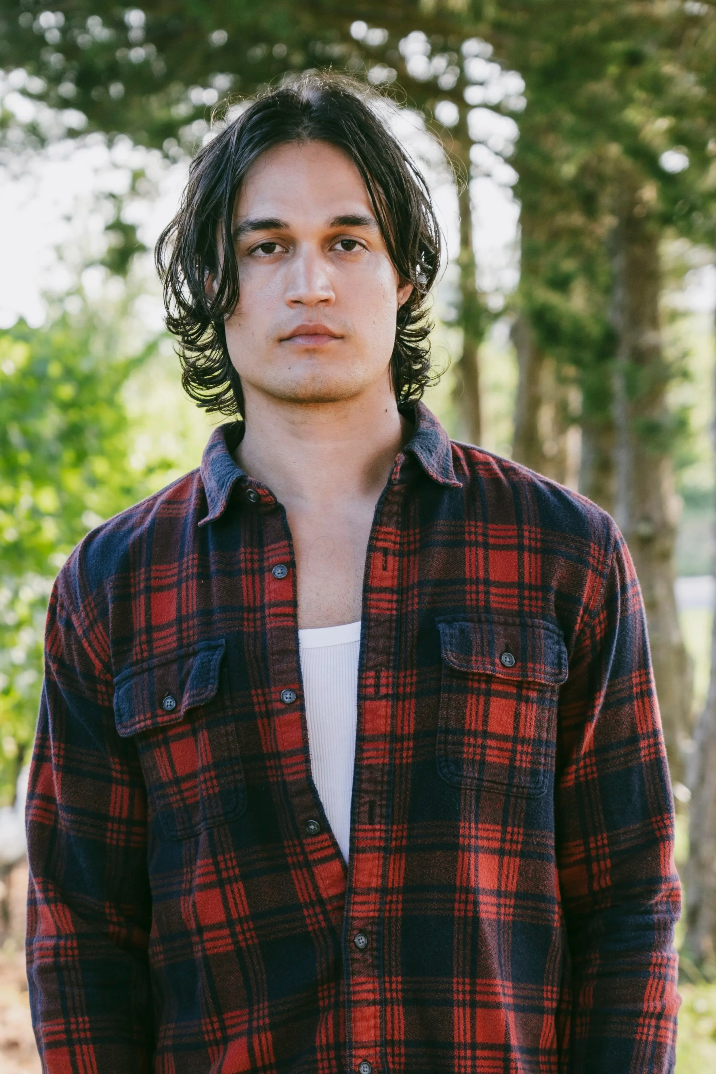 A young man with medium-length dark hair, wearing a red and black plaid shirt, stands outdoors with trees and a sunny sky in the background.