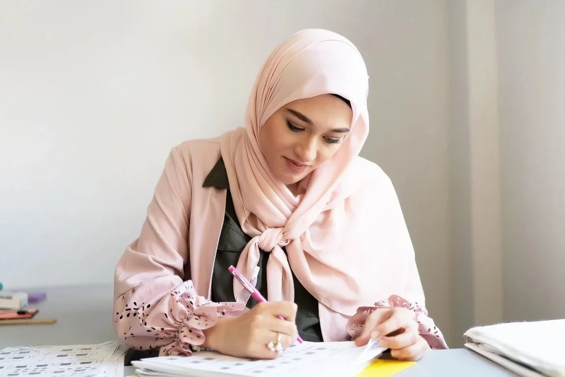 A woman wearing a pink hijab and a pink blouse sitting at a desk, writing in a notebook with loose papers and files around her.