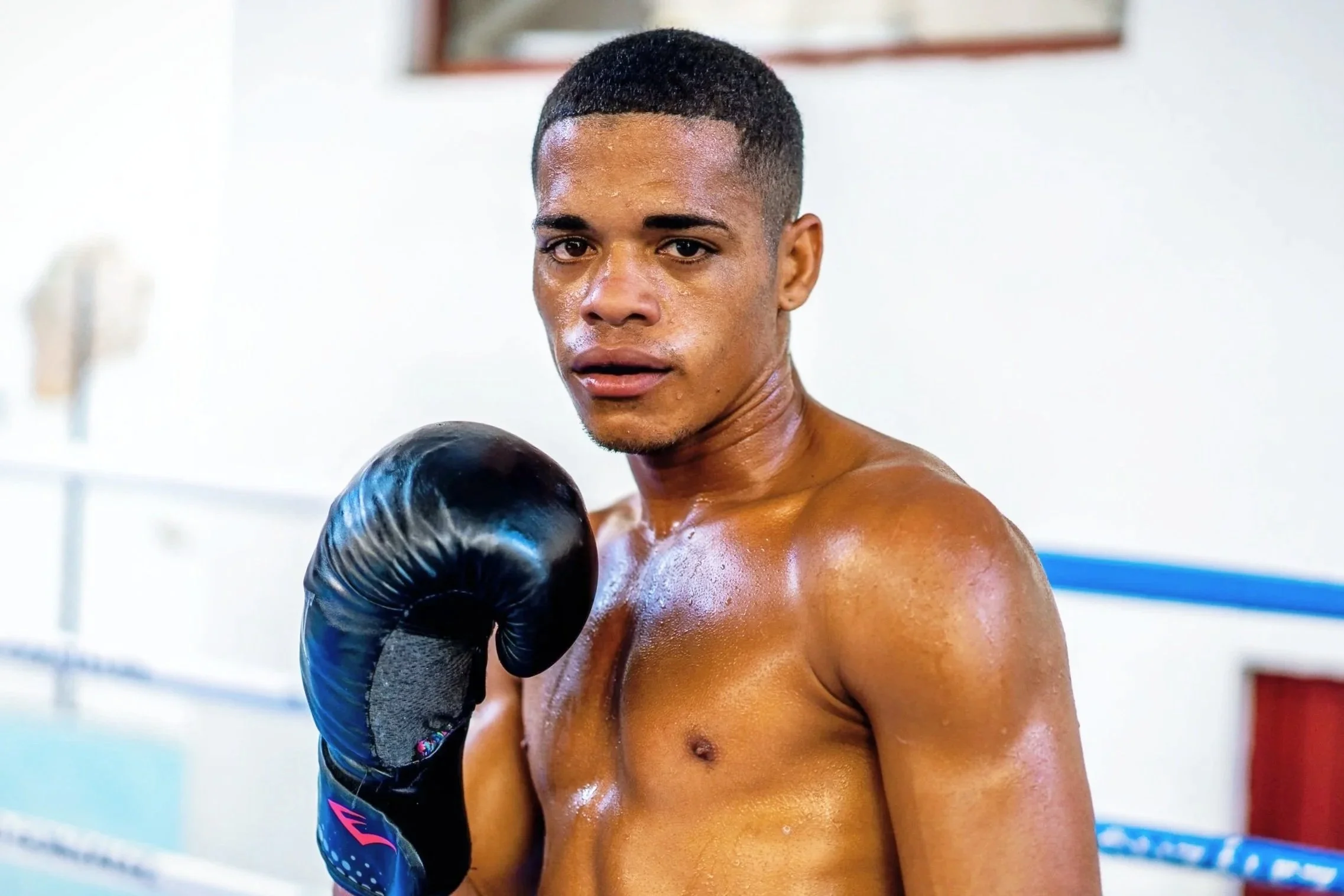 A shirtless male boxer with a sweat-glossed face and black boxing glove on his left hand, looking directly at the camera inside a boxing gym with white walls.
