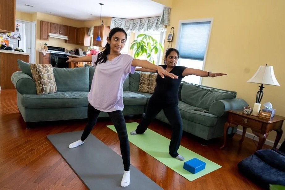 Two women practicing yoga indoors on mats, with arms extended and one leg slightly bent, in a living room with yellow walls, a green couch, lamp, side table, and kitchen in the background.