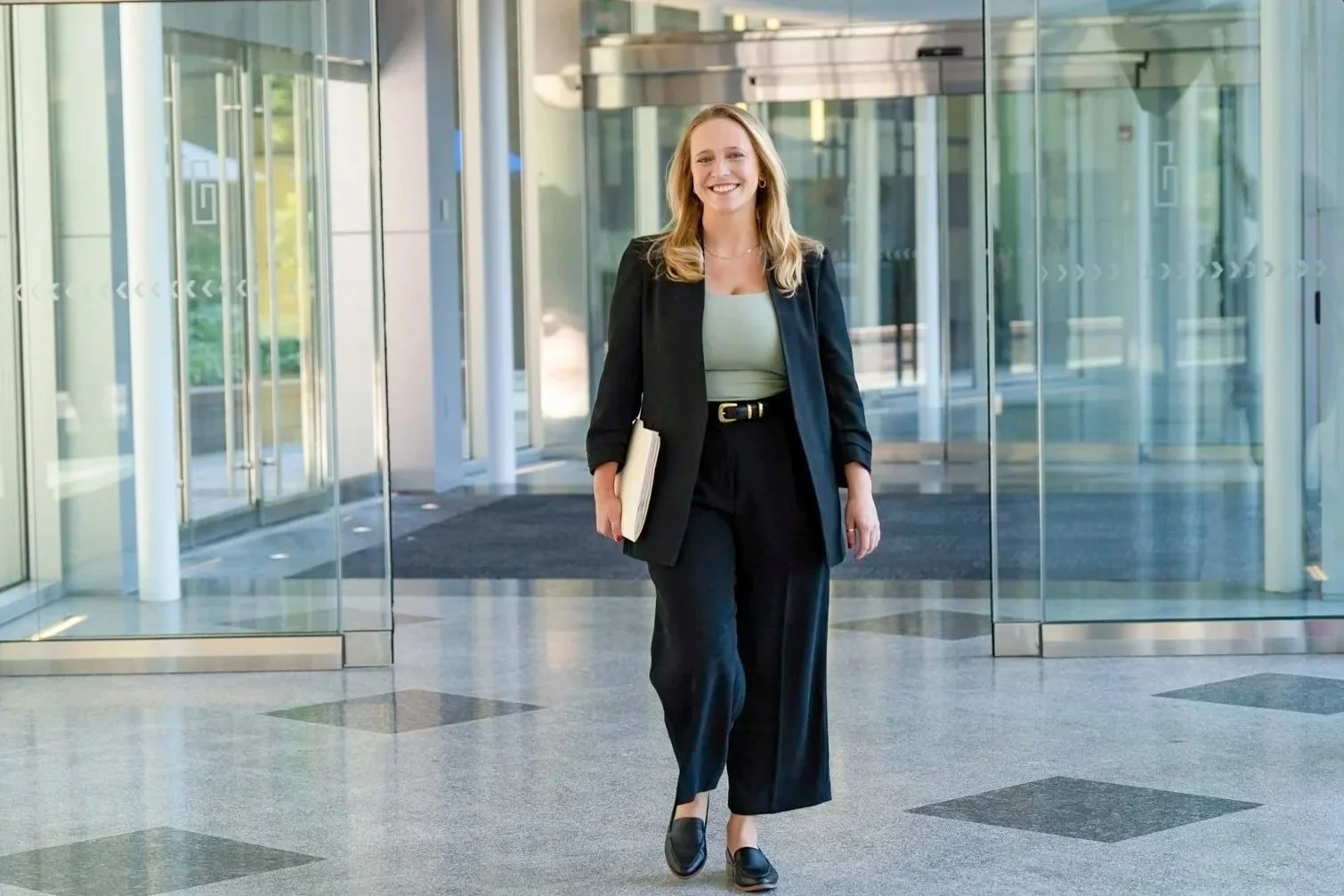 A professional woman in business attire walking out of a modern office building entrance holding a folder.