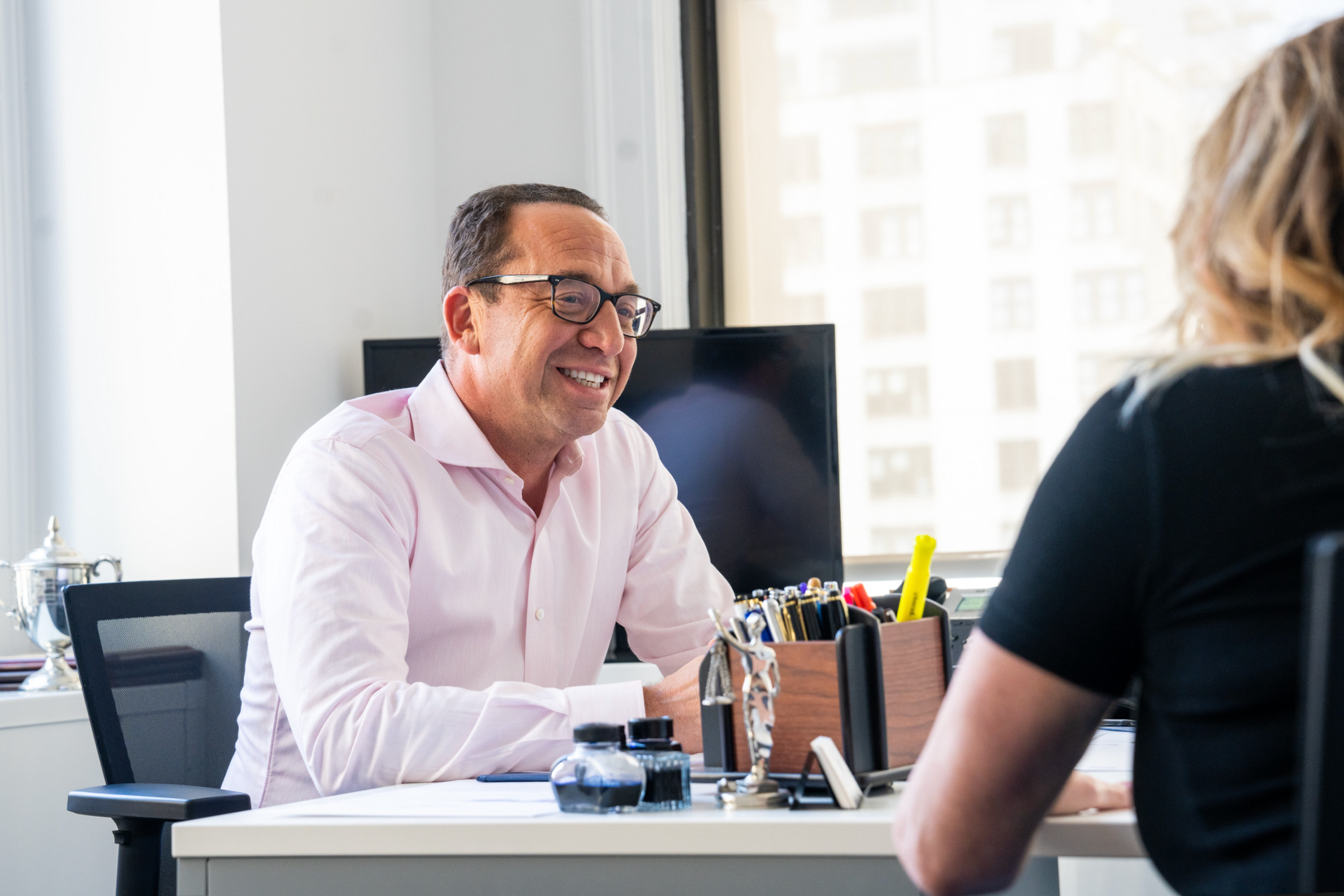 A man wearing glasses and a pink shirt smiling as he talks with a woman sitting across from him in an office. The office is bright, with large windows showing a cityscape, and the desk has various office supplies.