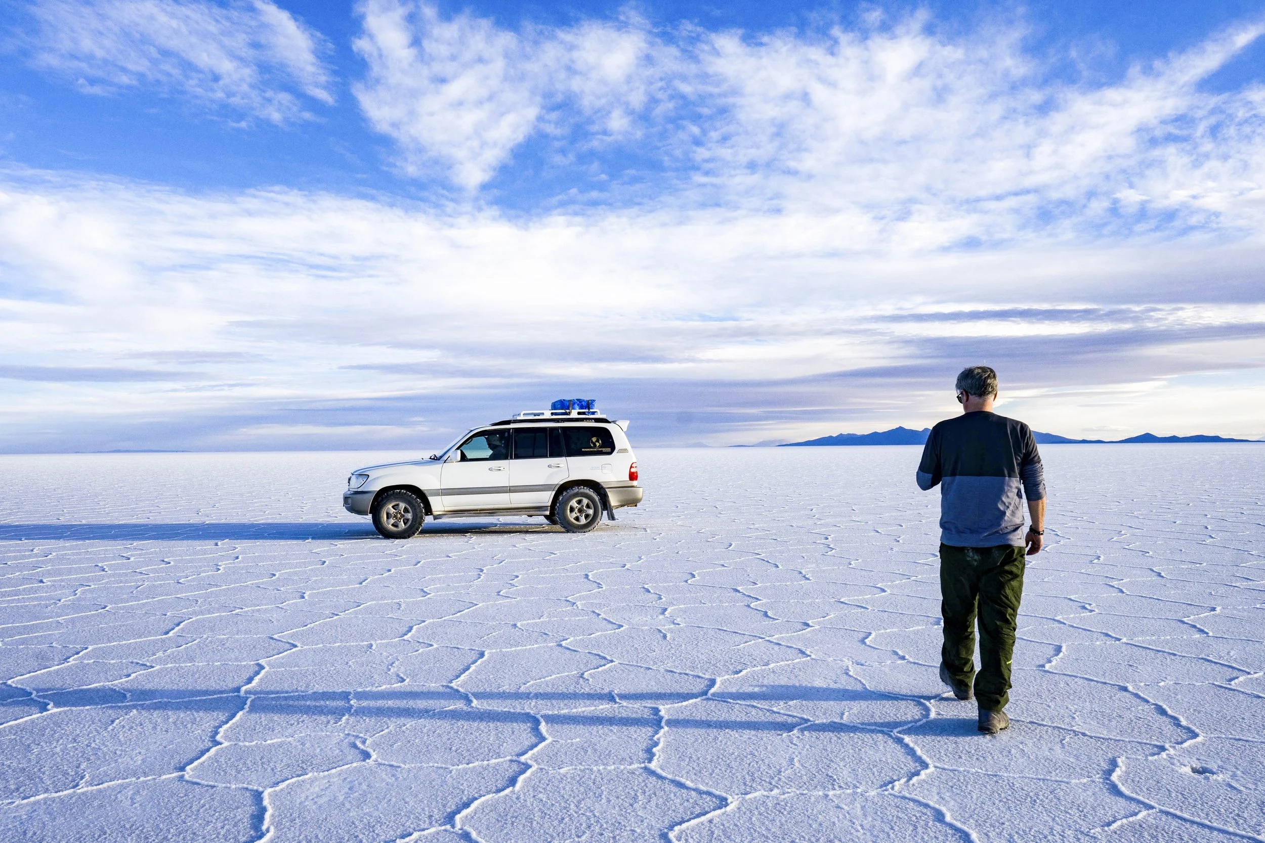 A person walking on a vast salt flat towards a parked white SUV with a blue roof rack, under a partly cloudy sky with mountains in the distant background.