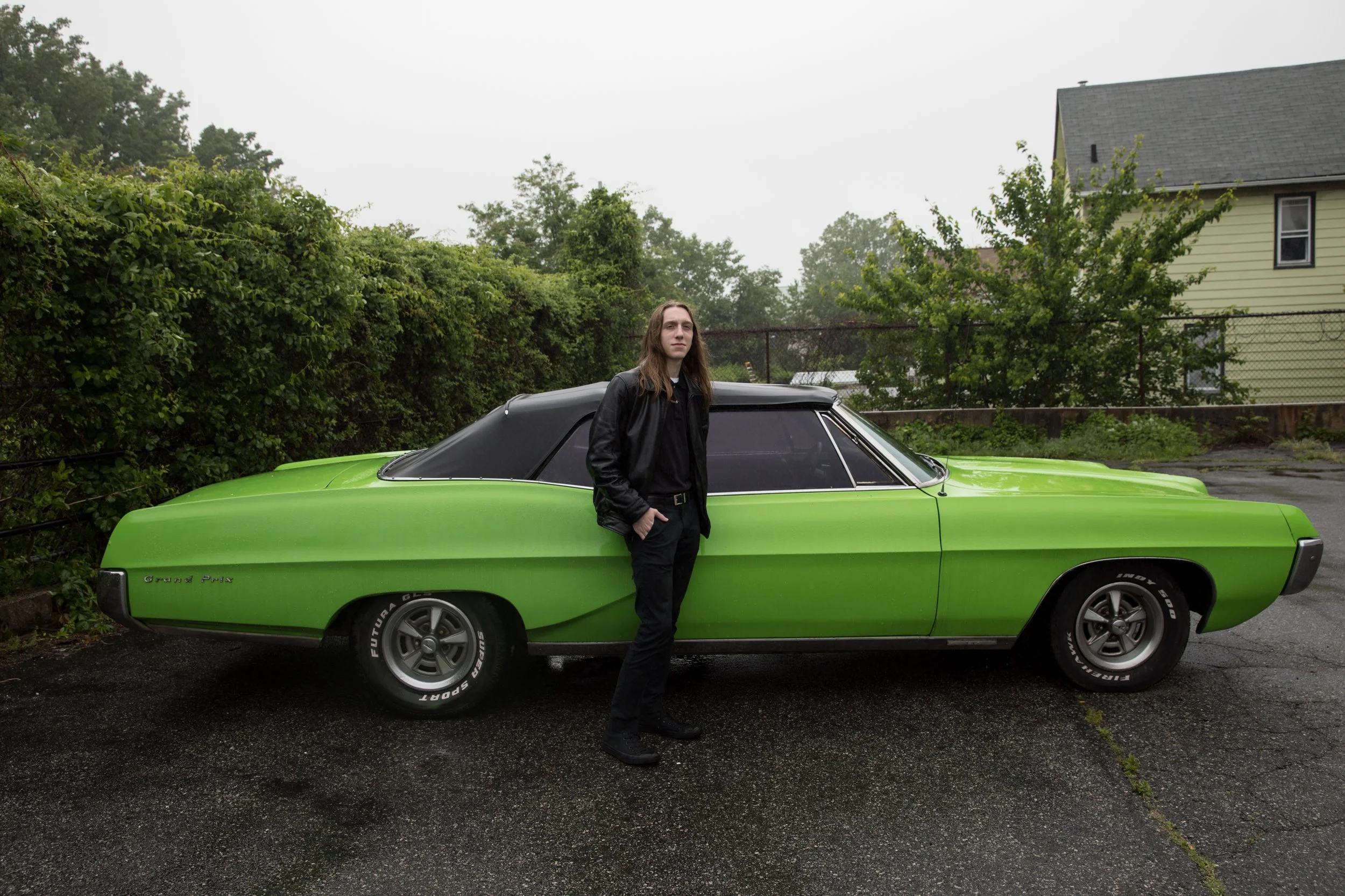 A young man with long hair, dressed in black, standing next to a bright green classic car with a black roof on an overcast day.