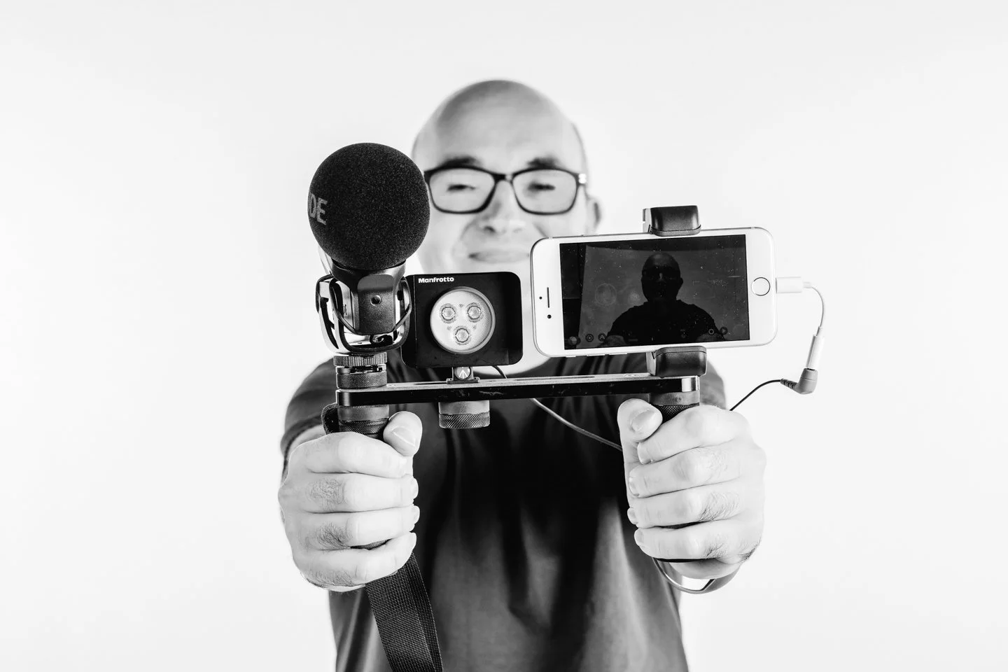 Man with glasses holding a camera rig with a microphone, a camera, and a smartphone in front of a plain white background.